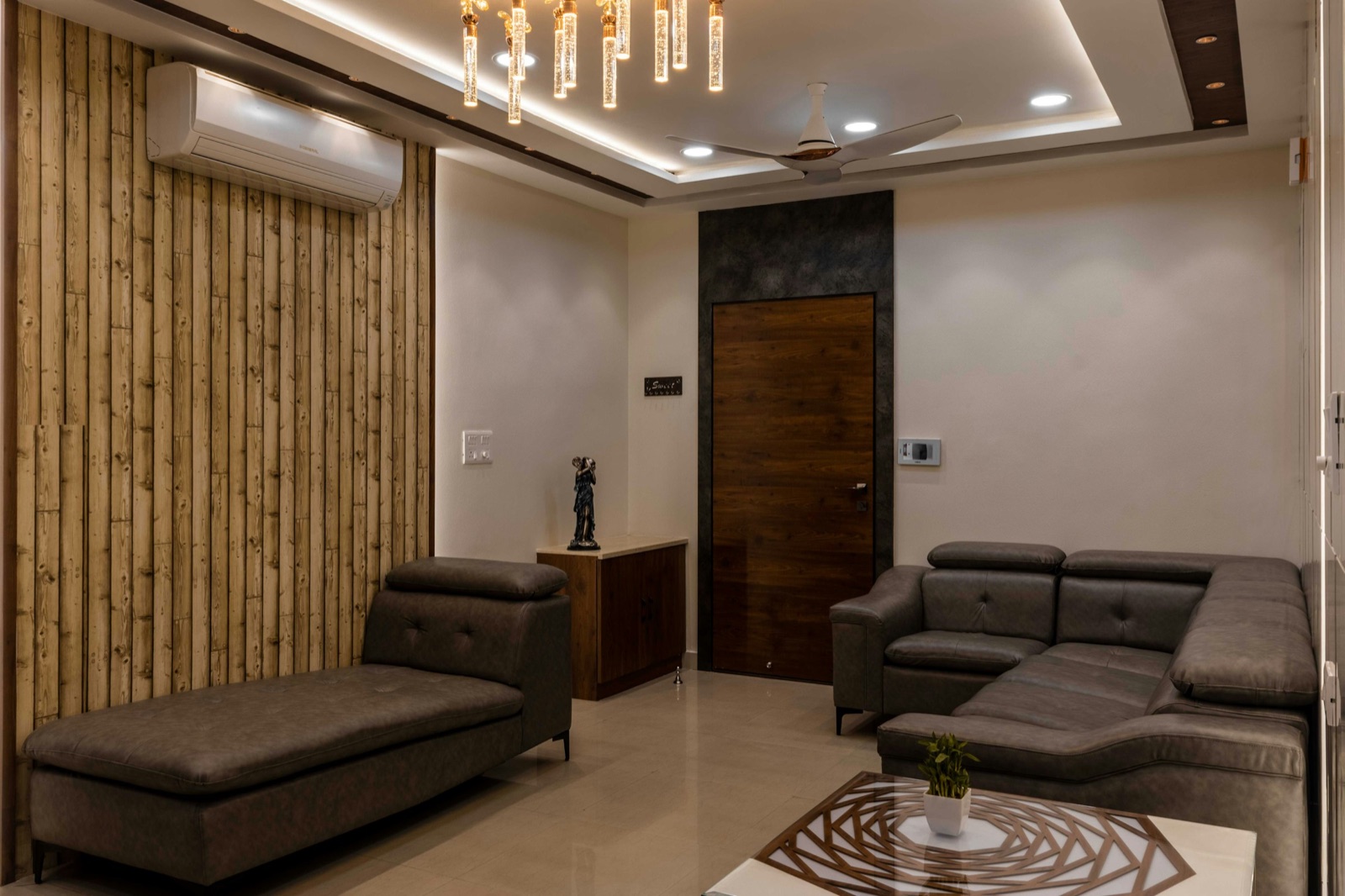 A Chennai flat's living room with a grey leatherette L-shaped sectional sofa with adjustable headrests in one corner, a separate matching chaise against a wood-textured wall, a white coffee table with a cut-work top, a split AC above, a crystal pendant light and the wooden main door between the two pieces