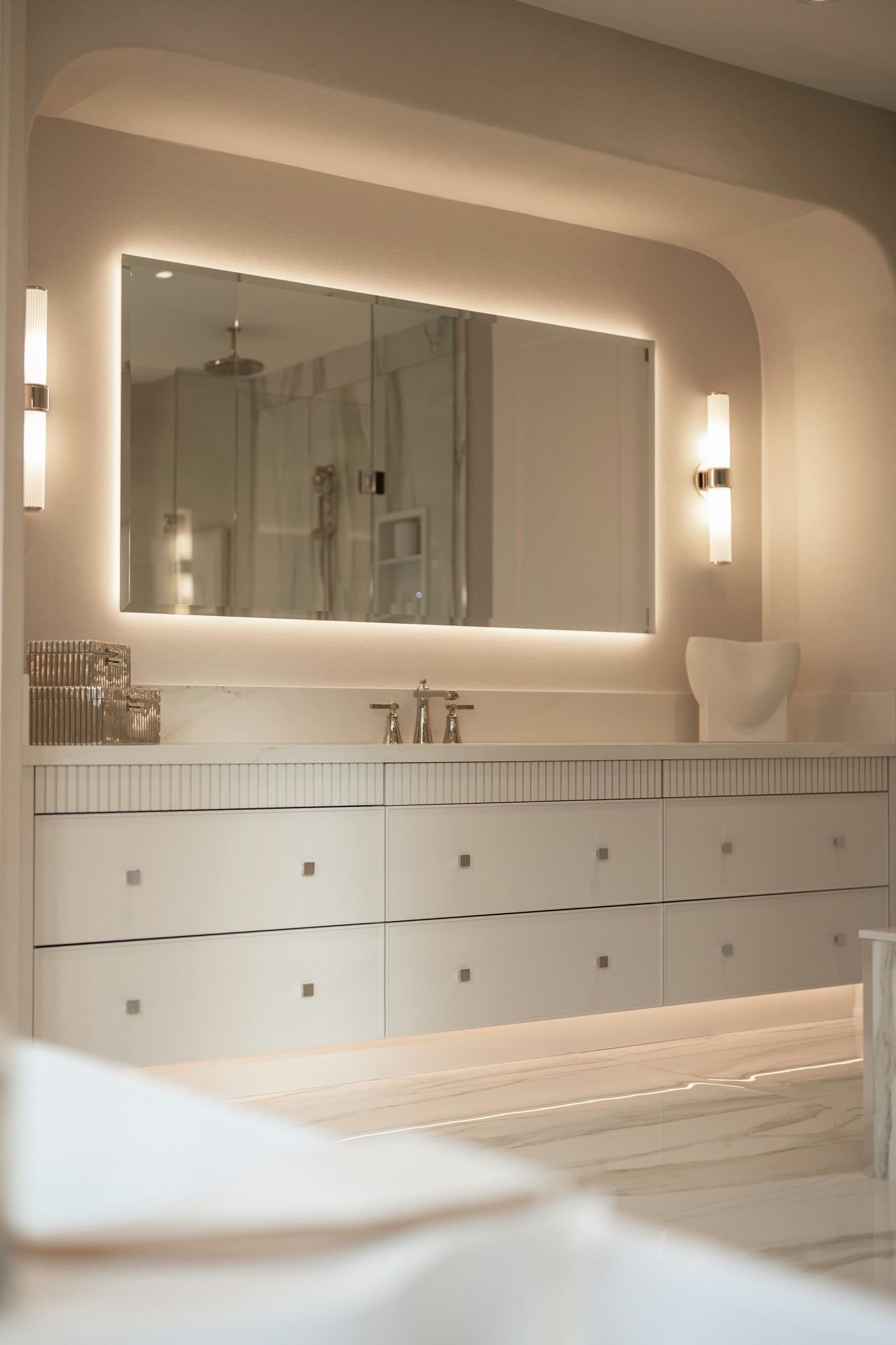 A long white wall-hung vanity with fluted drawer fronts and a marble top under a wide frameless mirror with a soft LED glow around its edges, flanked by two slim ribbed-glass wall lights, set in a curved recess; the mirror reflects a glass shower and marble wall.