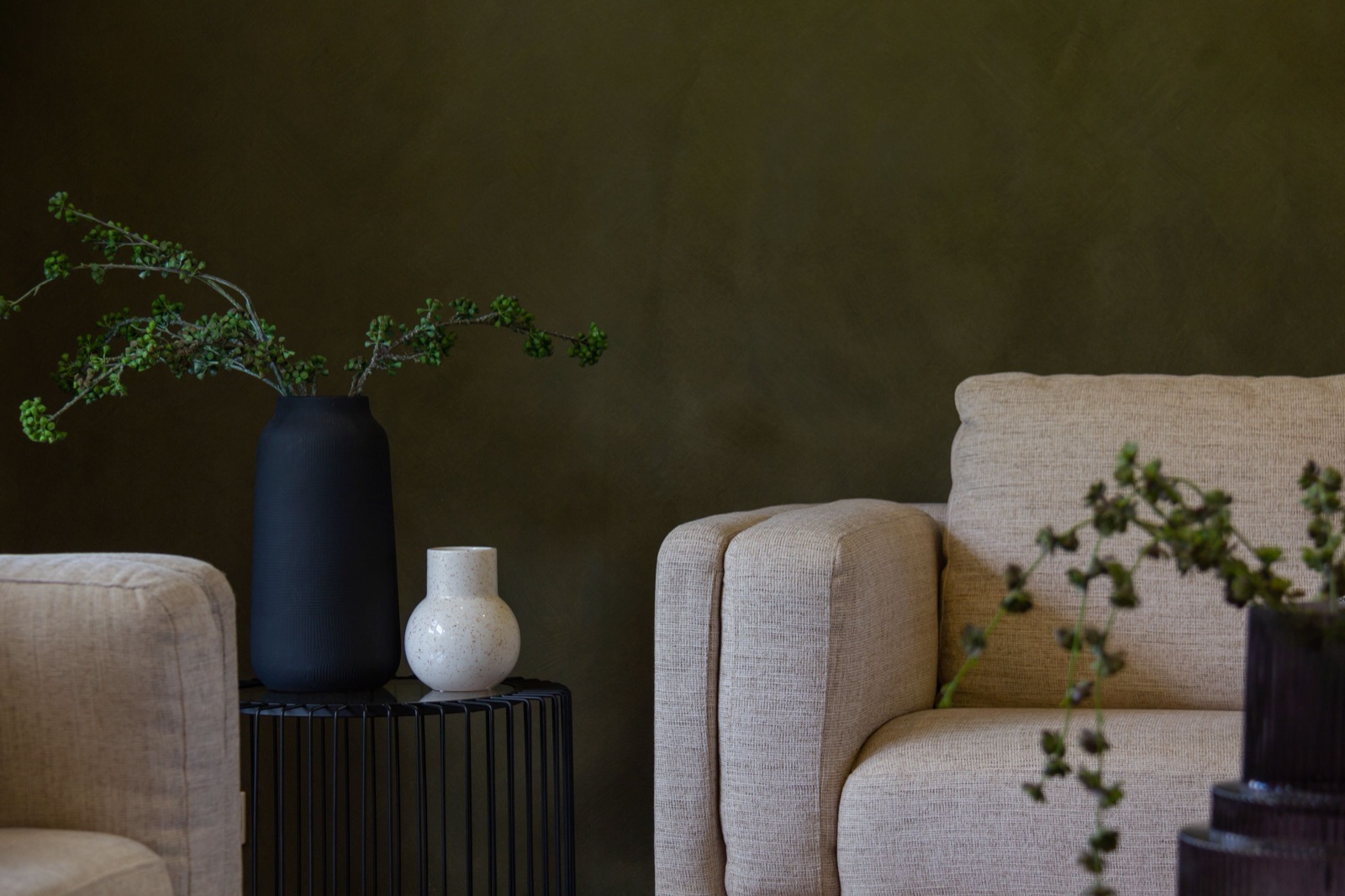 Living room with a deep olive green limewash-style wall, two oatmeal fabric armchairs, a black ribbed vase with green berry stems and a small white vase on a black wire side table