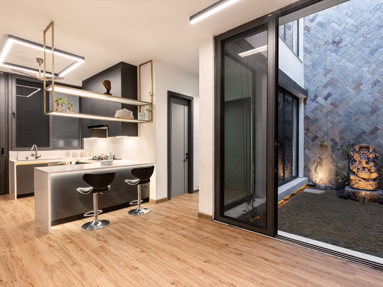 Open kitchen with dark handle-less cabinets, a white counter and waterfall peninsula with two bar stools, a brass-framed shelf hung from the ceiling, square linear ceiling lights, glow lighting under the counter edge, and a full-height glass door opening to a stone-clad courtyard.