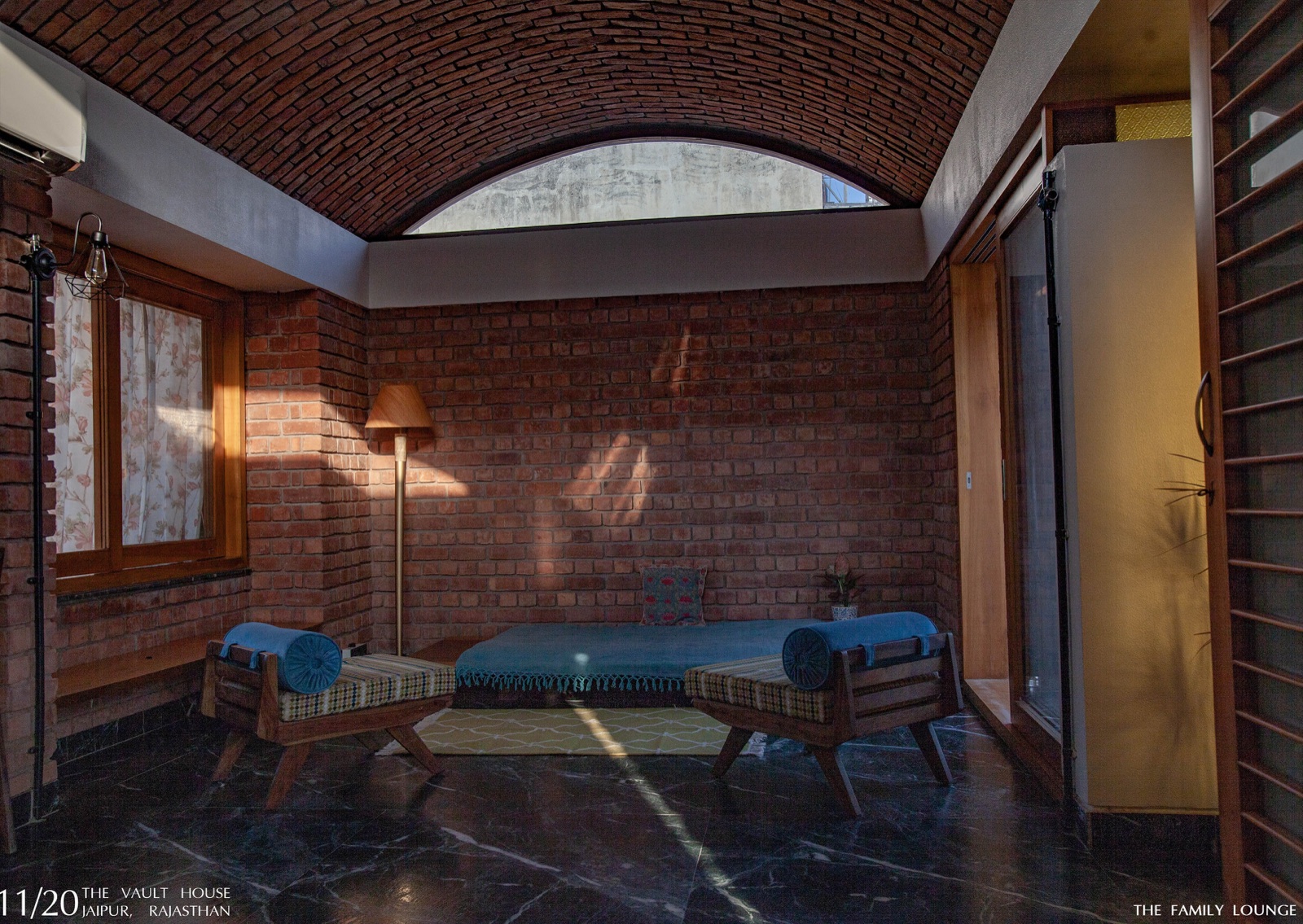 Family lounge in a Jaipur house with a brick barrel-vaulted ceiling, an arched clerestory window, exposed brick walls, a low daybed, two timber benches with blue bolsters and a dark marble floor