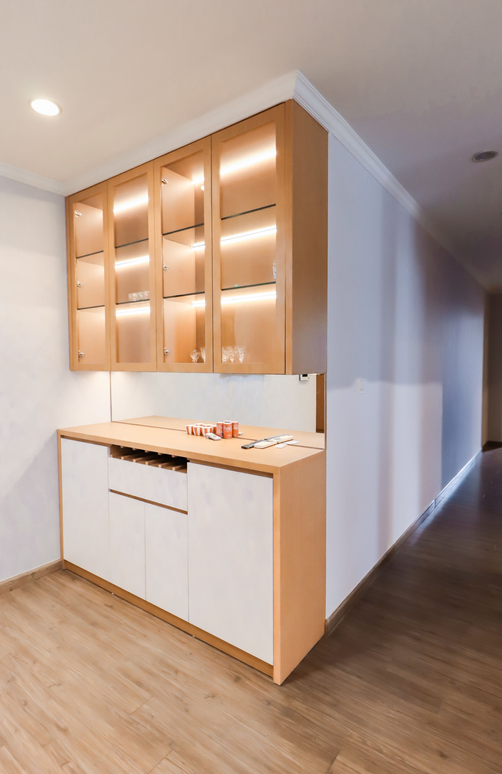 Crockery unit at the end of a hallway: four lit glass-fronted wall cabinets in light oak holding glasses, above a white and oak base cabinet with a wooden counter, a slatted bottle rack and a backsplash panel.
