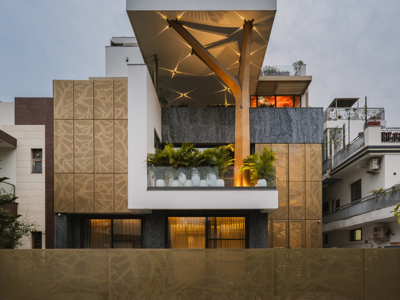 Front of a contemporary house in Panchkula at dusk: gold-toned perforated panels with a branch pattern, a white cantilevered terrace with glass railing and palms, dark textured stone below, and a matching perforated boundary wall along the street