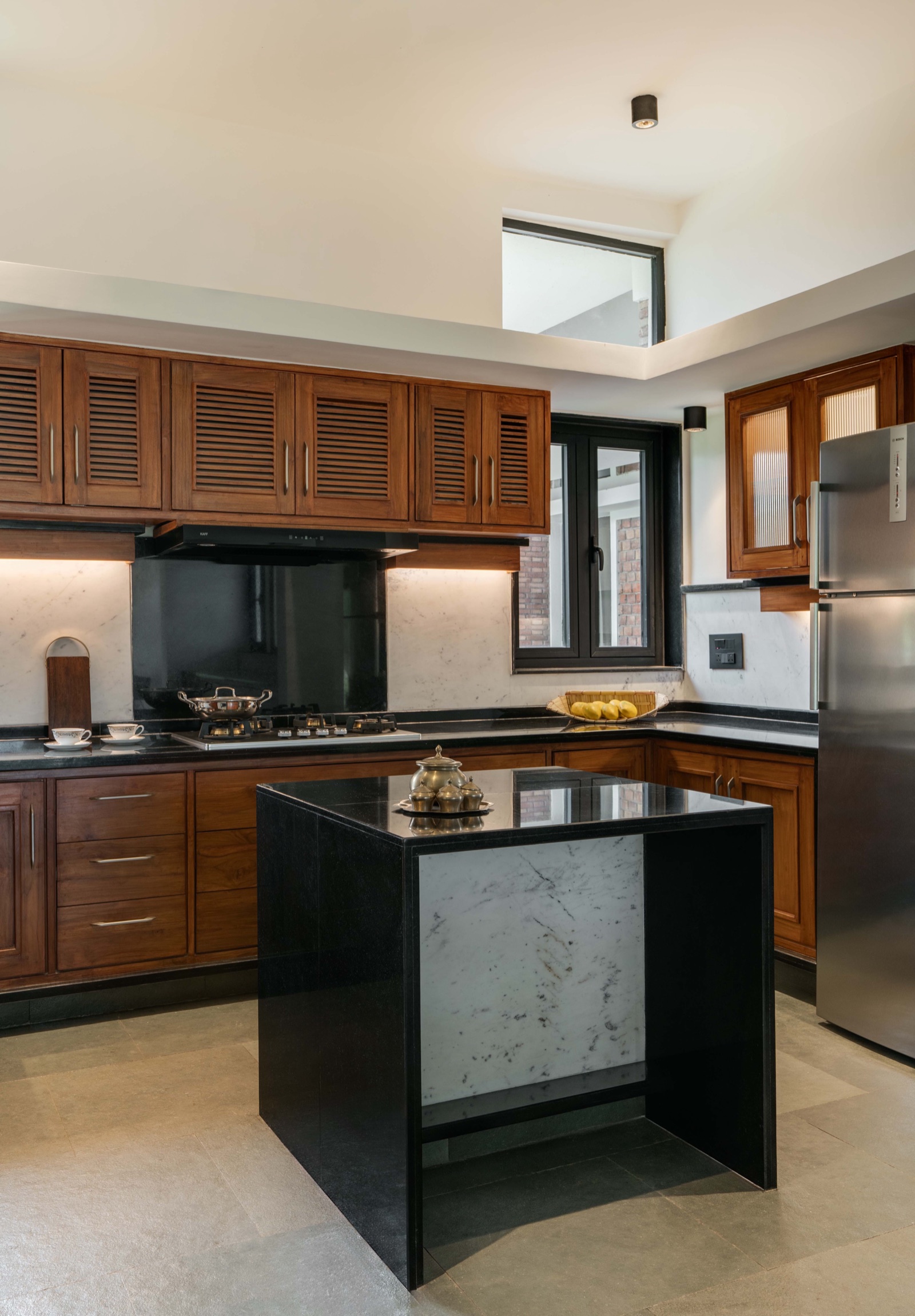 Kitchen with teak-toned louvred wall cabinets and panelled base drawers, a black stone counter, white marble backsplash with a black glass panel behind the gas hob and chimney, a black stone island with a white marble inset, a steel refrigerator and a high clerestory window.
