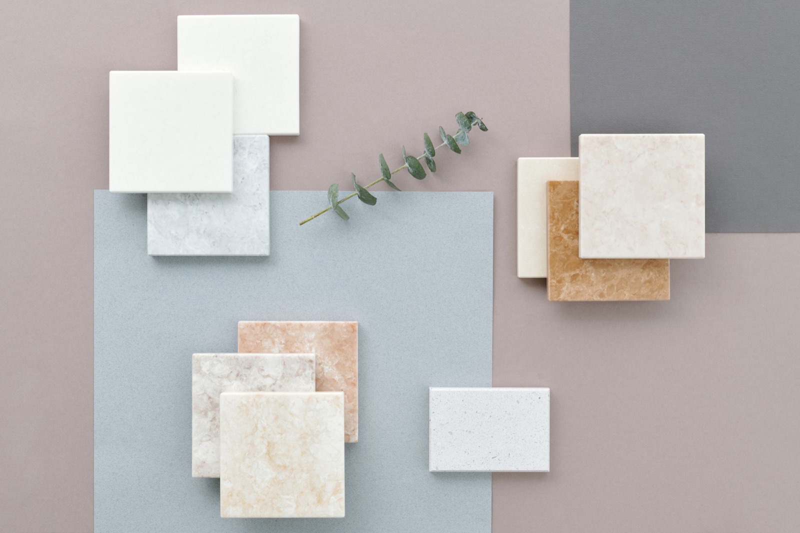 Flat lay of square stone-look samples in white, pale grey, cream, peach and amber marble patterns, grouped on grey and mauve card with a sprig of eucalyptus
