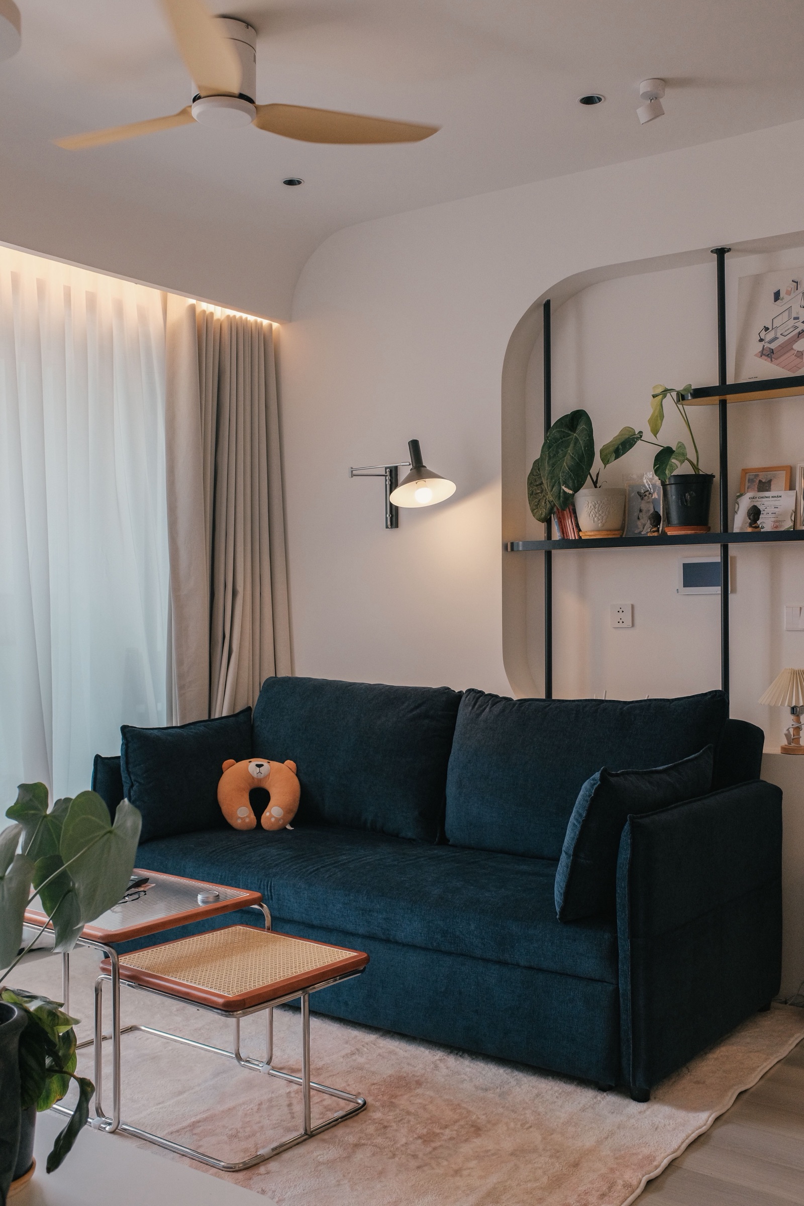 Living room with a flat white ceiling, a three-blade ceiling fan, small recessed downlights and a surface spotlight, and a dropped band with rounded ends along the window wall that hides the curtain track and washes the sheer and linen curtains with warm cove light, above a navy sofa and nesting side tables.