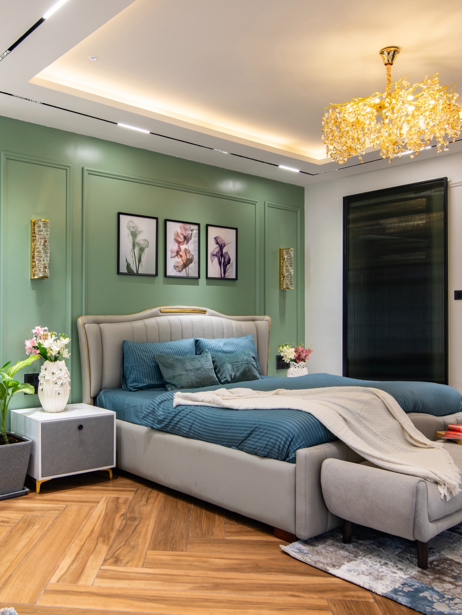 Photograph of a master bedroom with a sage green bed wall finished in raised moulding panels, three framed floral prints and two gold wall lights above a grey upholstered bed, a cove-lit false ceiling and herringbone wood-look flooring.