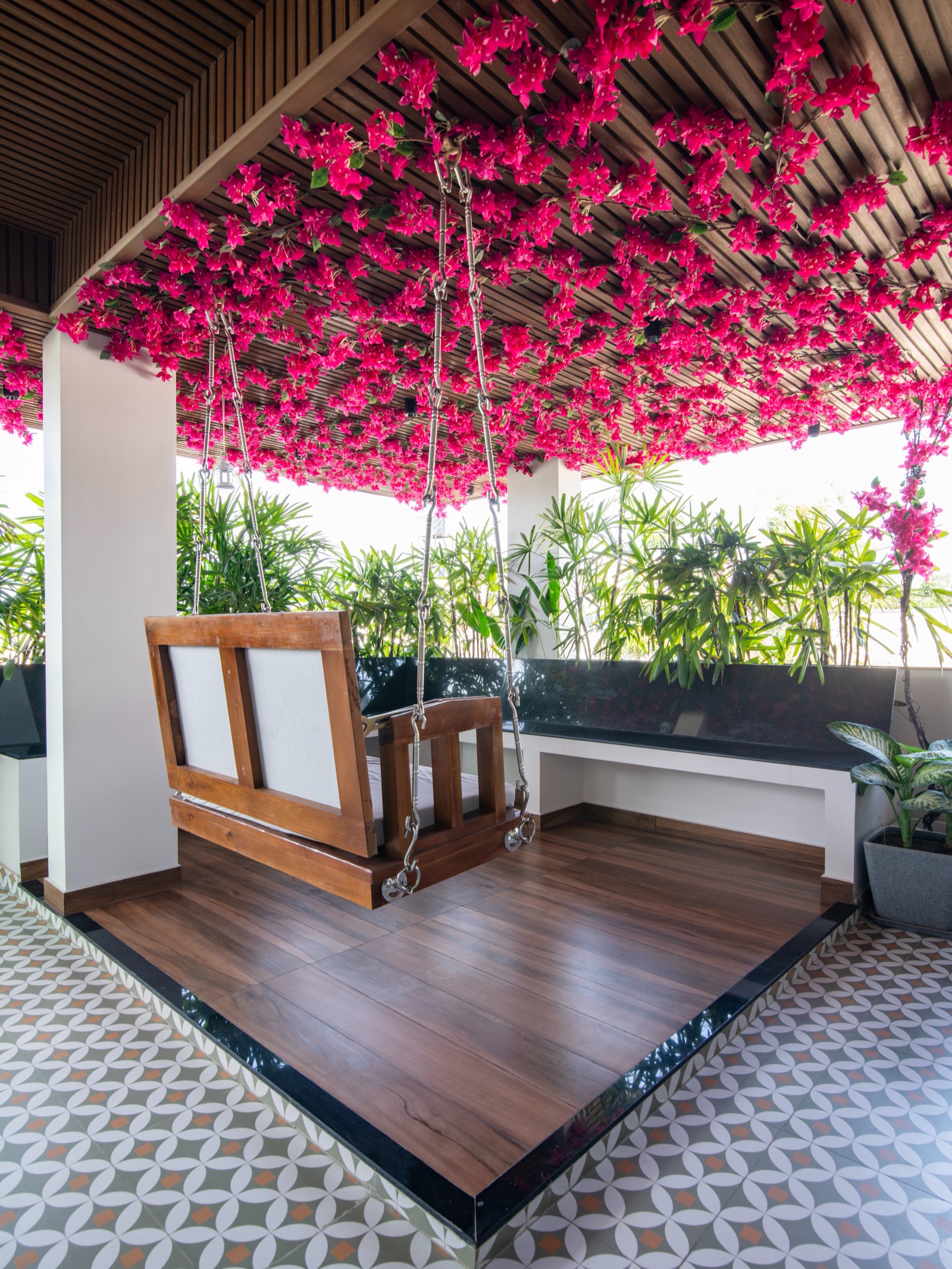 A covered rooftop terrace with a wooden swing over a raised platform finished in wood-look planks edged in black stone, surrounded by a floor of grey, white and terracotta patterned tiles, with potted palms along a black stone ledge and pink bougainvillea across a slatted ceiling