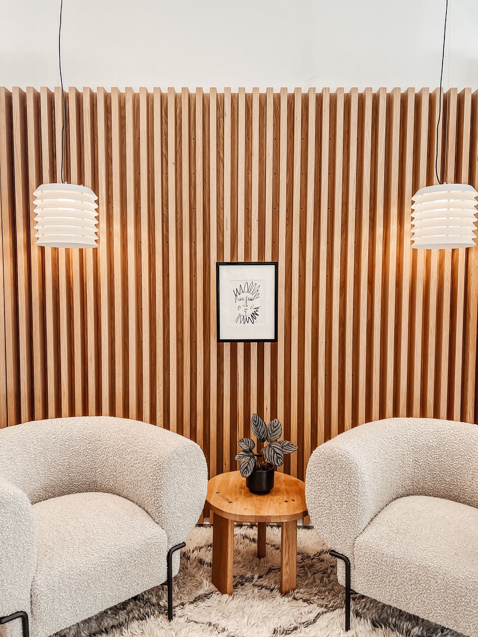 Photograph of a wall of light wood slats with gaps between them and small fixings visible on the face of each slat, two white tiered pendant lamps, a small framed sketch, two cream boucle armchairs and a round wooden side table on a shaggy rug.