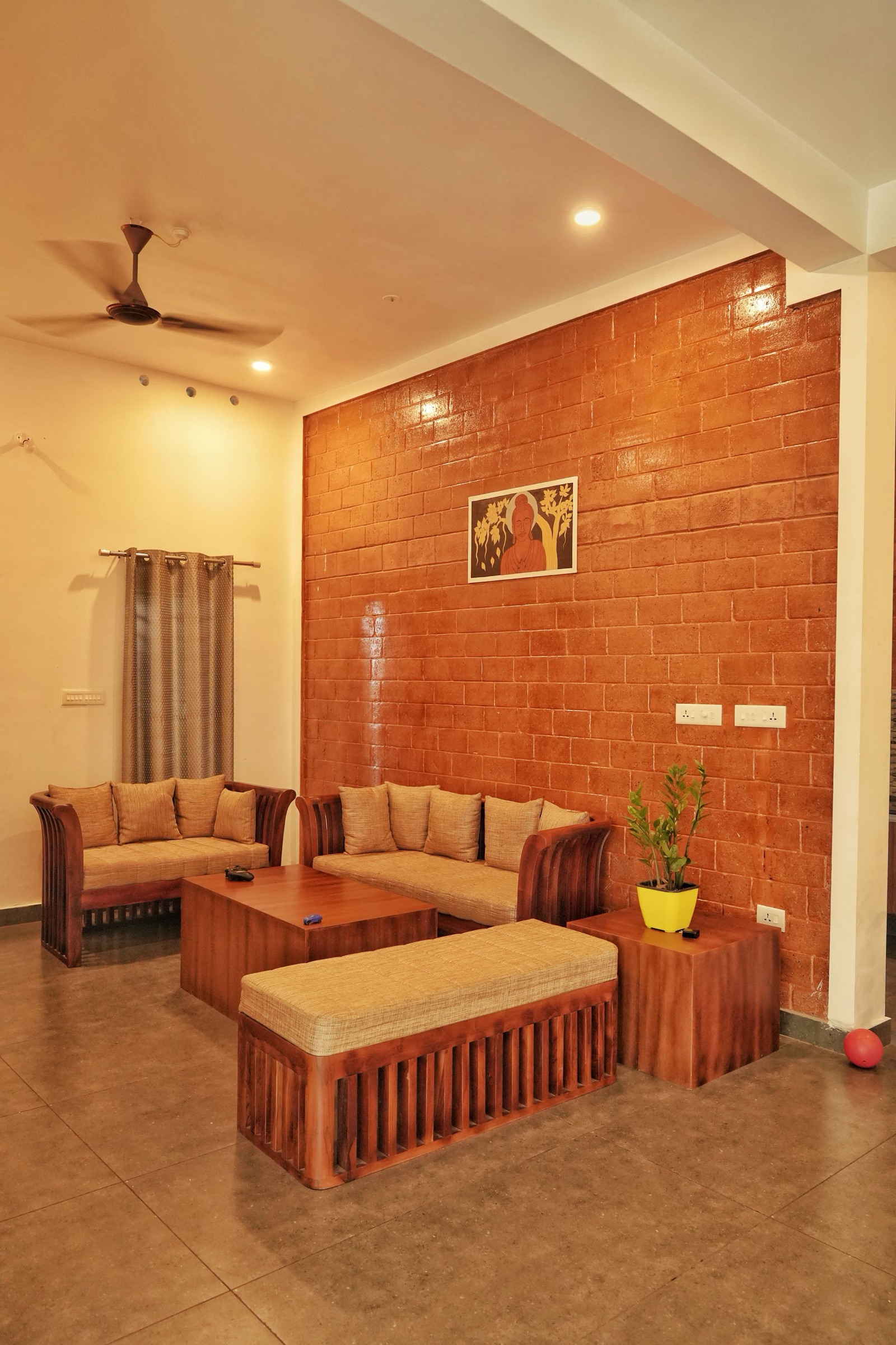 Living room in a Kerala home with a textured brick accent wall, two slatted wooden sofas with beige woven cushions, a square wooden coffee table, a cushioned wooden bench, a side table with a ZZ plant in a yellow pot, a grey curtain on a rod and a ceiling fan