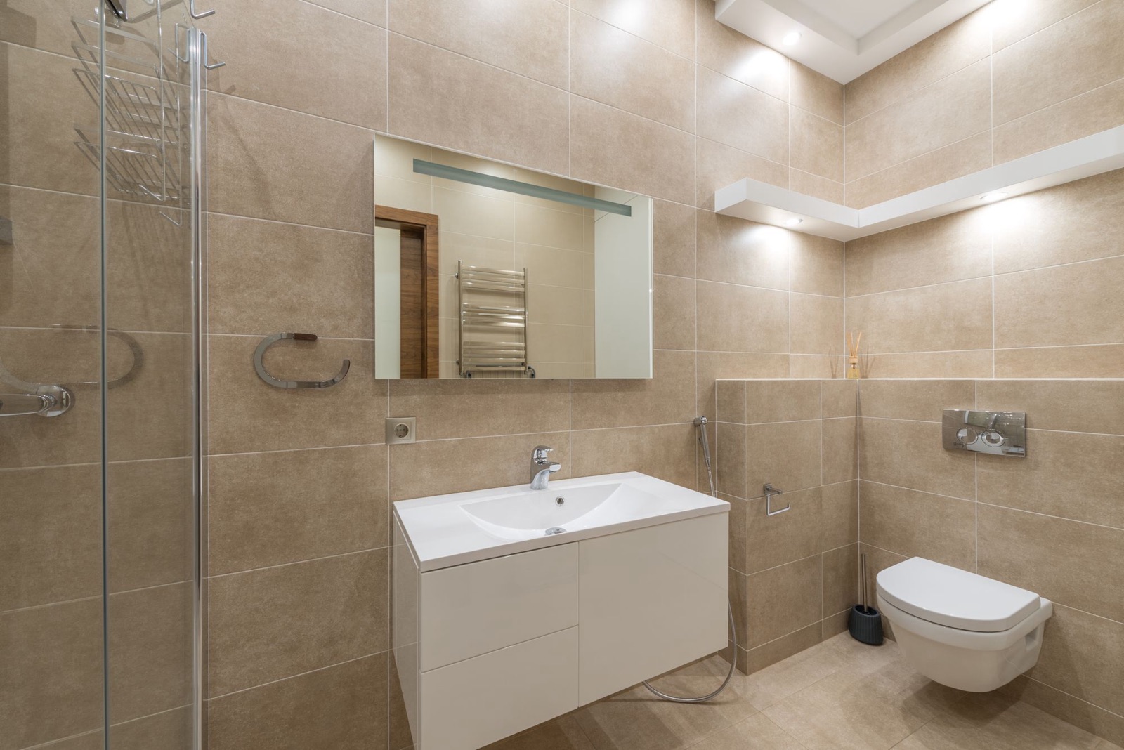 A compact bathroom tiled floor to ceiling in beige matt rectangular tiles laid in a stack, with a glass shower screen, a white wall-hung vanity, a mirror with a light strip, a health faucet and a wall-hung WC