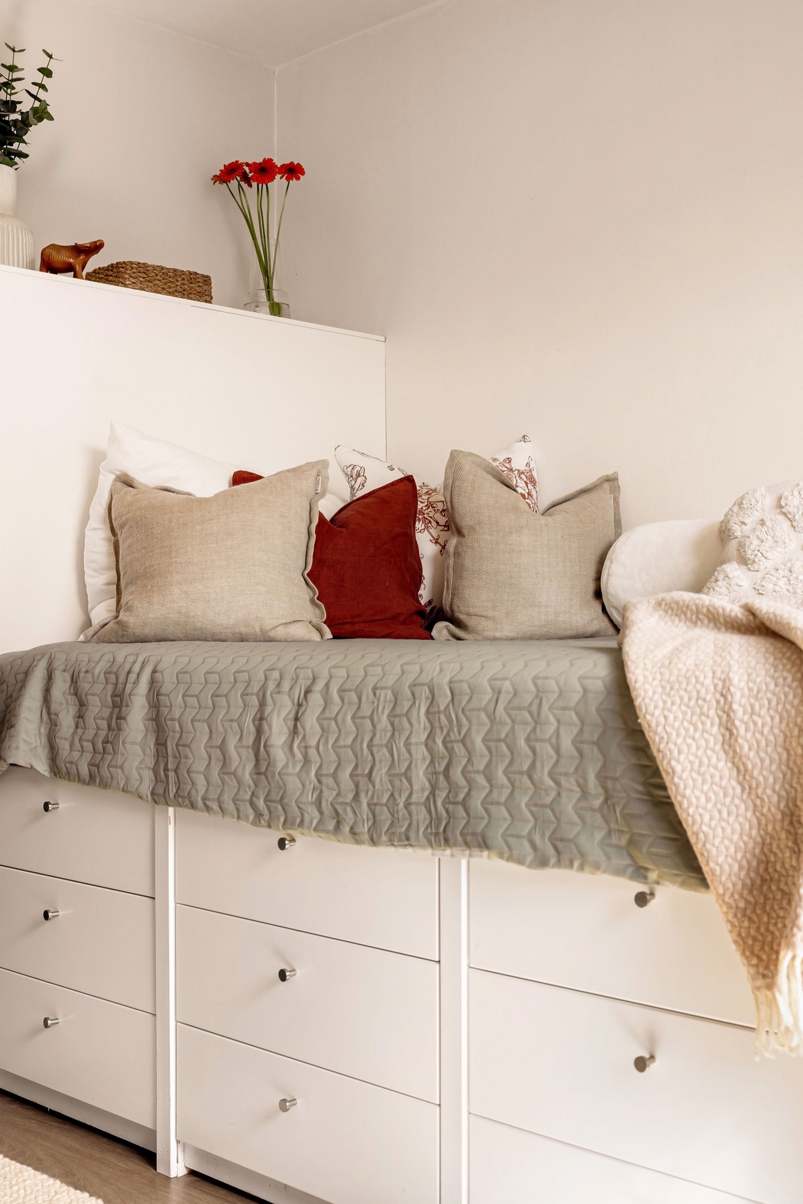 A raised single bed built on top of white chests of drawers with round knobs, made up with a grey quilt, linen and red cushions and a cream throw, against a pale wall