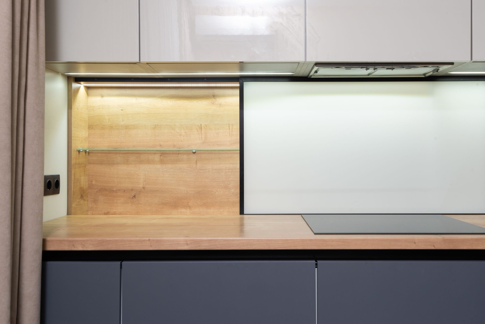 Close view of a kitchen run with glossy beige wall cabinets whose underside holds a slim built-in extractor hood above a black induction hob, an oak worktop and splashback panel with a glass shelf, a white glass splashback behind the hob and dark grey handle-less base drawers.