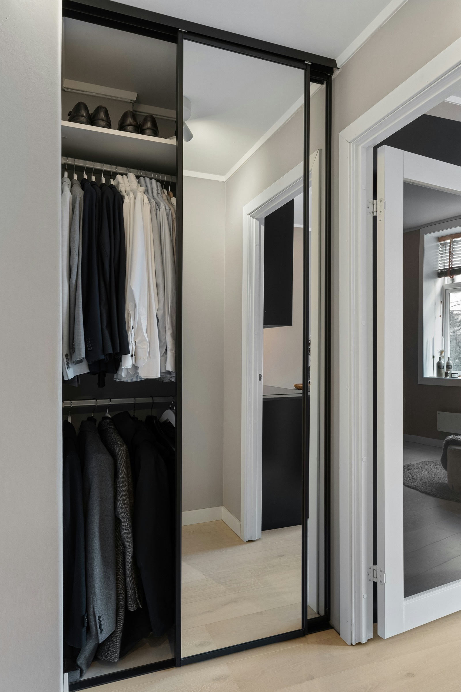 A built-in wardrobe with two black-framed sliding doors, one a full-height mirror and the other slid aside to show a top shelf of shoes and two hanging rails of shirts, jackets and coats.