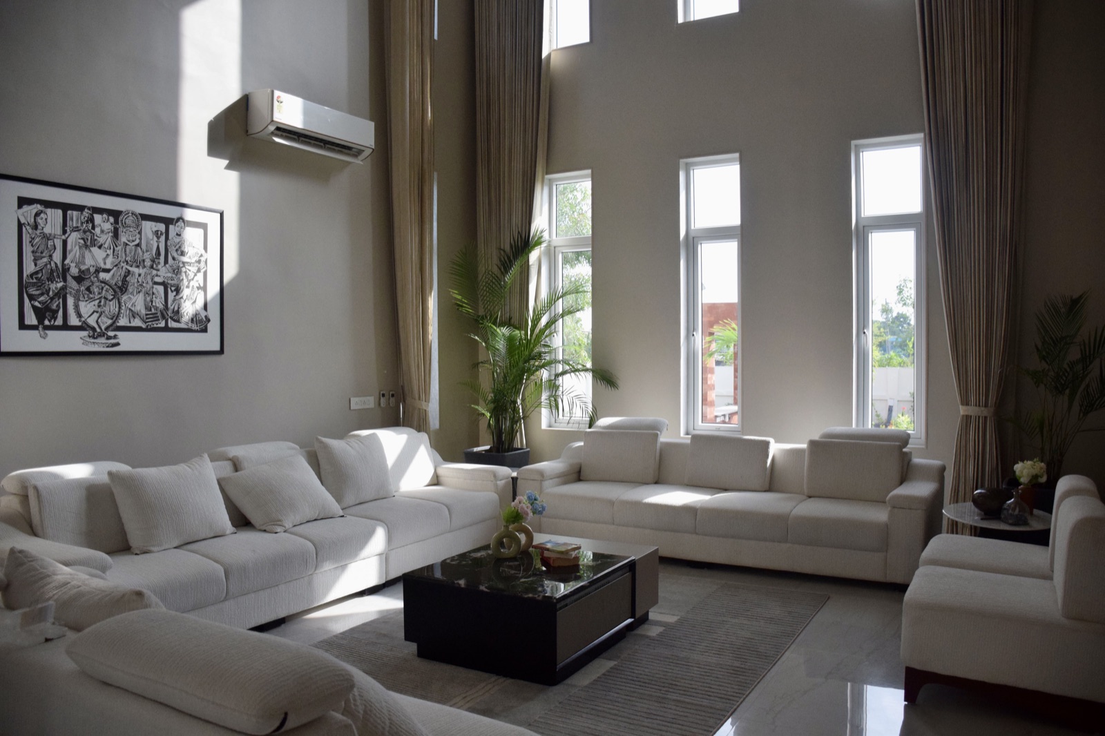 Double-height living room with white sofas around a black stone coffee table, tall curtained windows looking out to the garden and a black and white artwork on the wall