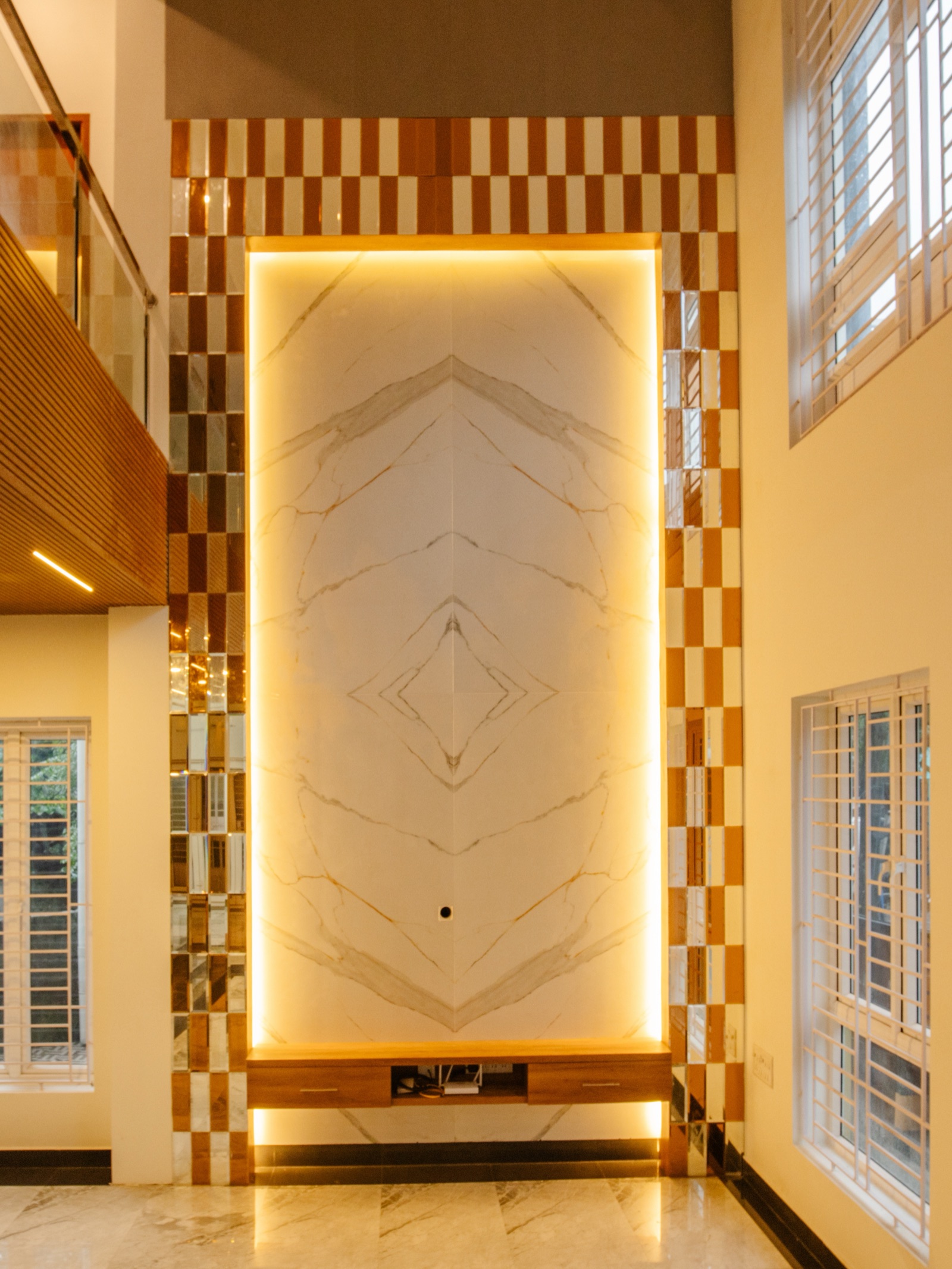 Double-height living room wall clad in bookmatched white marble with warm backlighting, framed by a checkerboard border of timber and mirror tiles, with a floating timber console below and tall windows with white grilles on either side