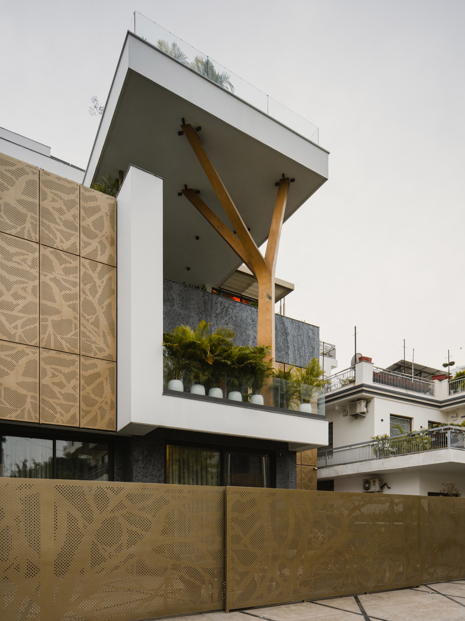 Side view of a contemporary Panchkula house: tall bronze-finish perforated metal panels with a leaf-vein pattern clad the upper floor and continue as the boundary screen in the foreground, beside a white cantilevered terrace with a glass railing, potted palms and a Y-shaped timber-finish column