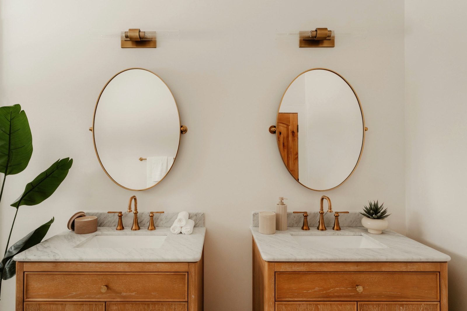 Two oak washstands with white marble tops and brass taps side by side, each under its own oval brass-framed pivot mirror narrower than the washstand, with a brass wall light centred above each mirror and a large leafy plant at the left.
