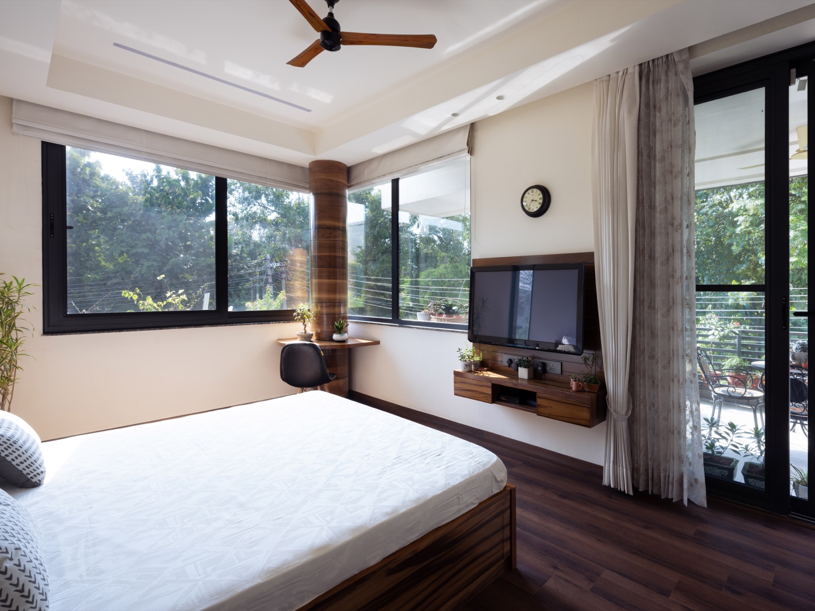 Bedroom with a white stepped false ceiling: a raised central tray with a long linear air-conditioning slot, a dropped band around the edge with small recessed downlights, and a three-blade wooden-finish ceiling fan hung in the tray. Corner windows with roller blinds look onto trees, a wall-mounted TV sits on a wood panel, and a glass door opens to a terrace.