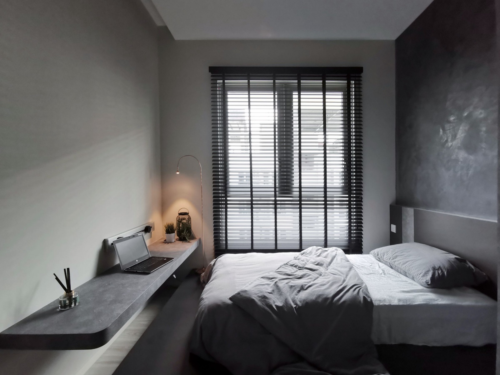 A narrow grey bedroom with a long wall-mounted concrete-look desk shelf running along one wall to the window, a laptop and a plant on it, a power track with a plug-in socket fixed to the wall just above the desk, a lantern lamp near the window with black venetian blinds, and a bed with grey bedding beside it.