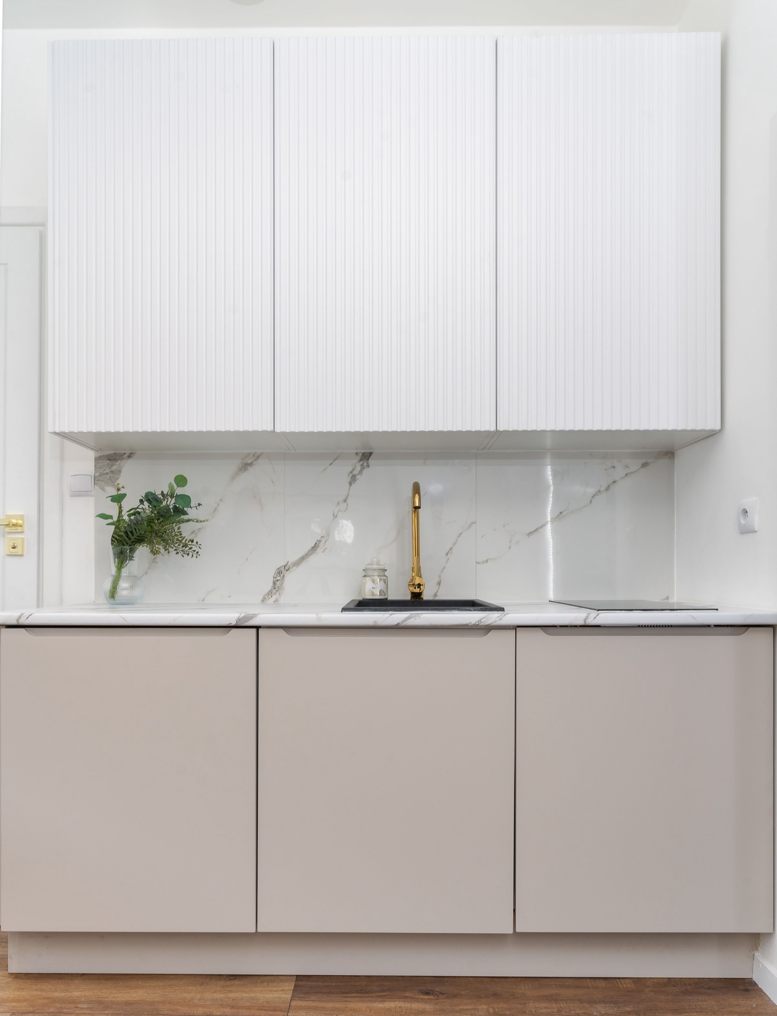 Straight kitchen with white fluted wall cabinets above taupe handle-less base units. Between the marble-look counter and the wall cabinets, three large glossy white panels with grey veining form the backsplash, with only two vertical joints, behind a black sink, a brass tap and a flush induction hob.