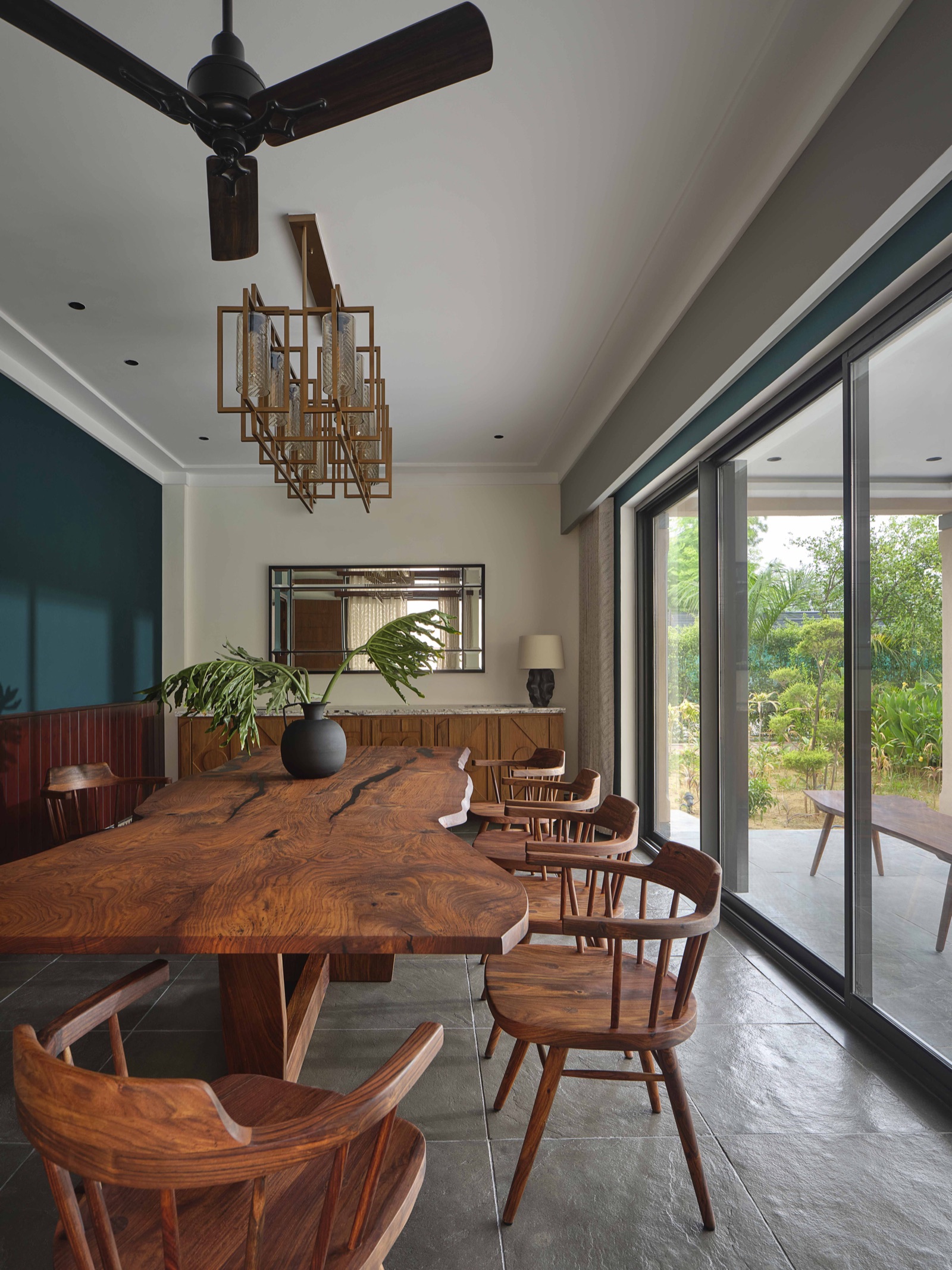 A long live-edge solid wood dining table with natural dark cracks running through the top, set on a wooden base with wooden spindle-back armchairs around it. A brass-framed linear pendant with ribbed glass shades hangs over the table, a carved wooden sideboard with a stone top sits under a mirror at the far end, the walls are teal above dark timber panelling, the floor is grey stone and full-height sliding glass doors open to a garden.
