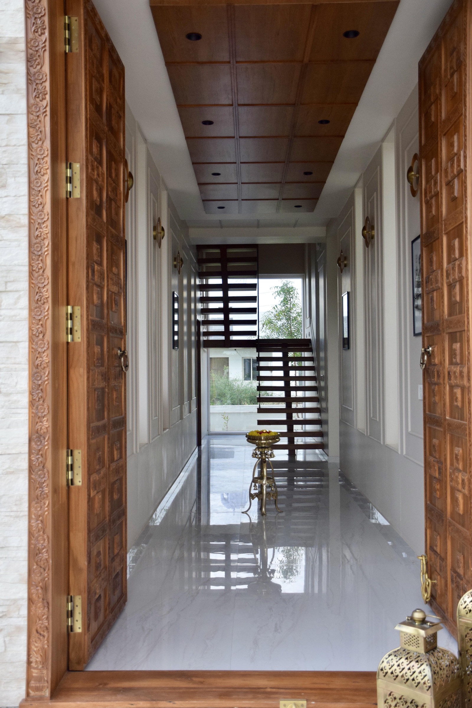 A pair of tall wooden main doors with deep carved panels and a carved floral frame, opened inward onto a white marble passage with a wood-panelled ceiling, brass wall lights and an open-riser staircase at the far end