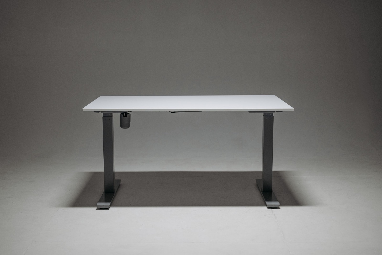 A white height-adjustable desk on a dark grey two-leg steel frame, photographed side-on against a grey studio backdrop, with a small motor housing under the top.
