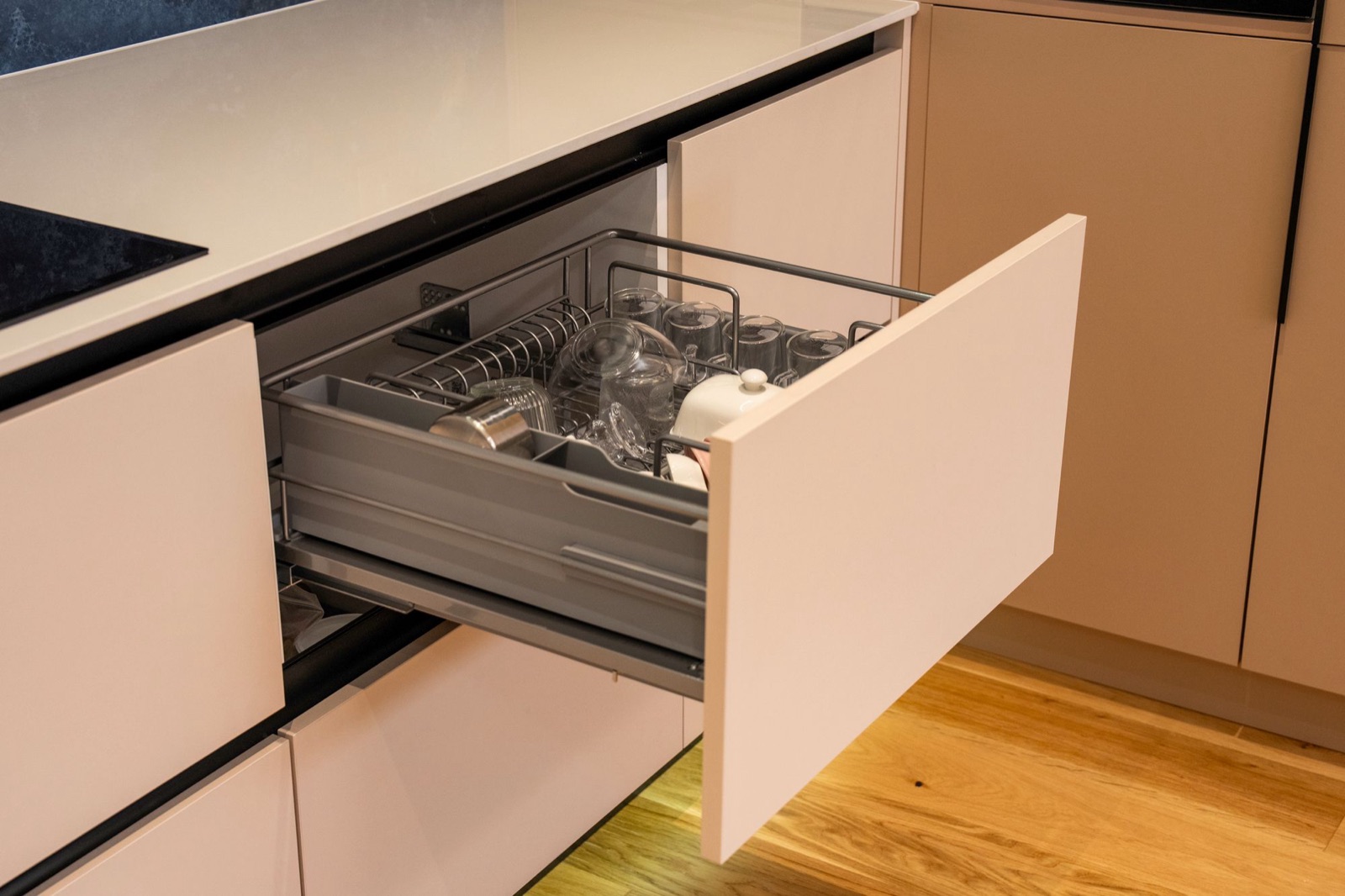A wide base-unit drawer pulled out to show a grey metal basket insert holding glasses, cups and a teapot, below a white countertop