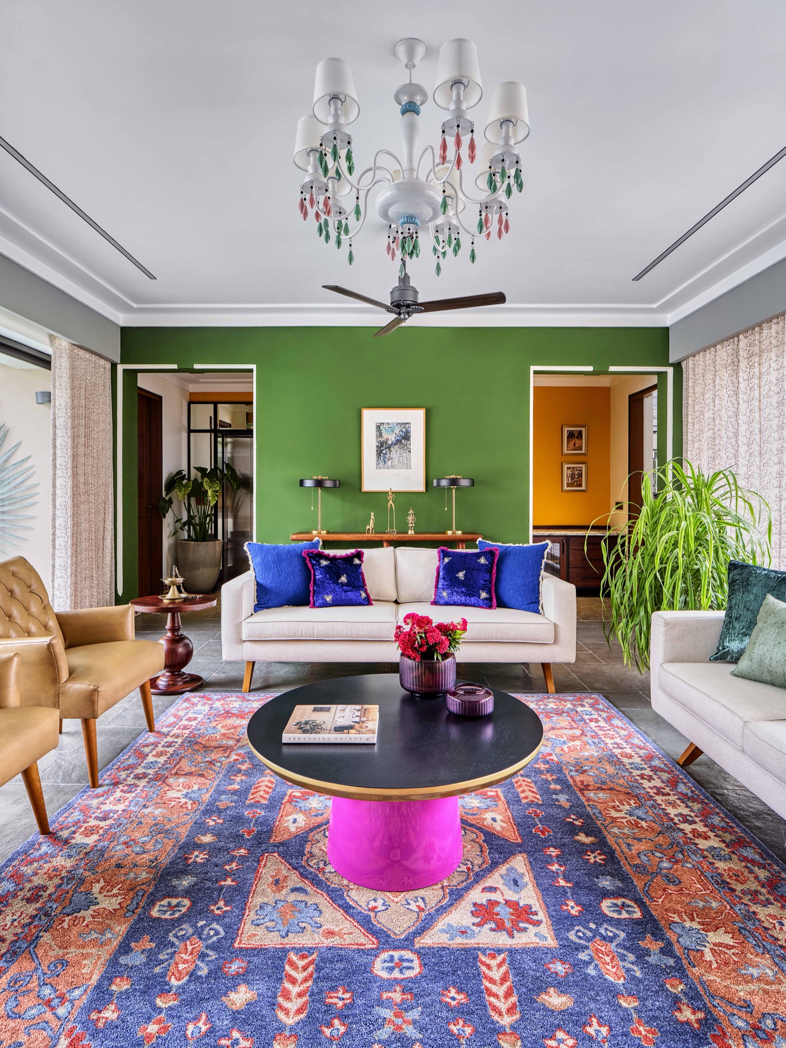 Living room with one deep green wall between two white-framed doorways, a white ceiling with cornice, cream sofas with royal blue cushions, a blue and rust patterned rug, and an ochre wall visible through the right doorway