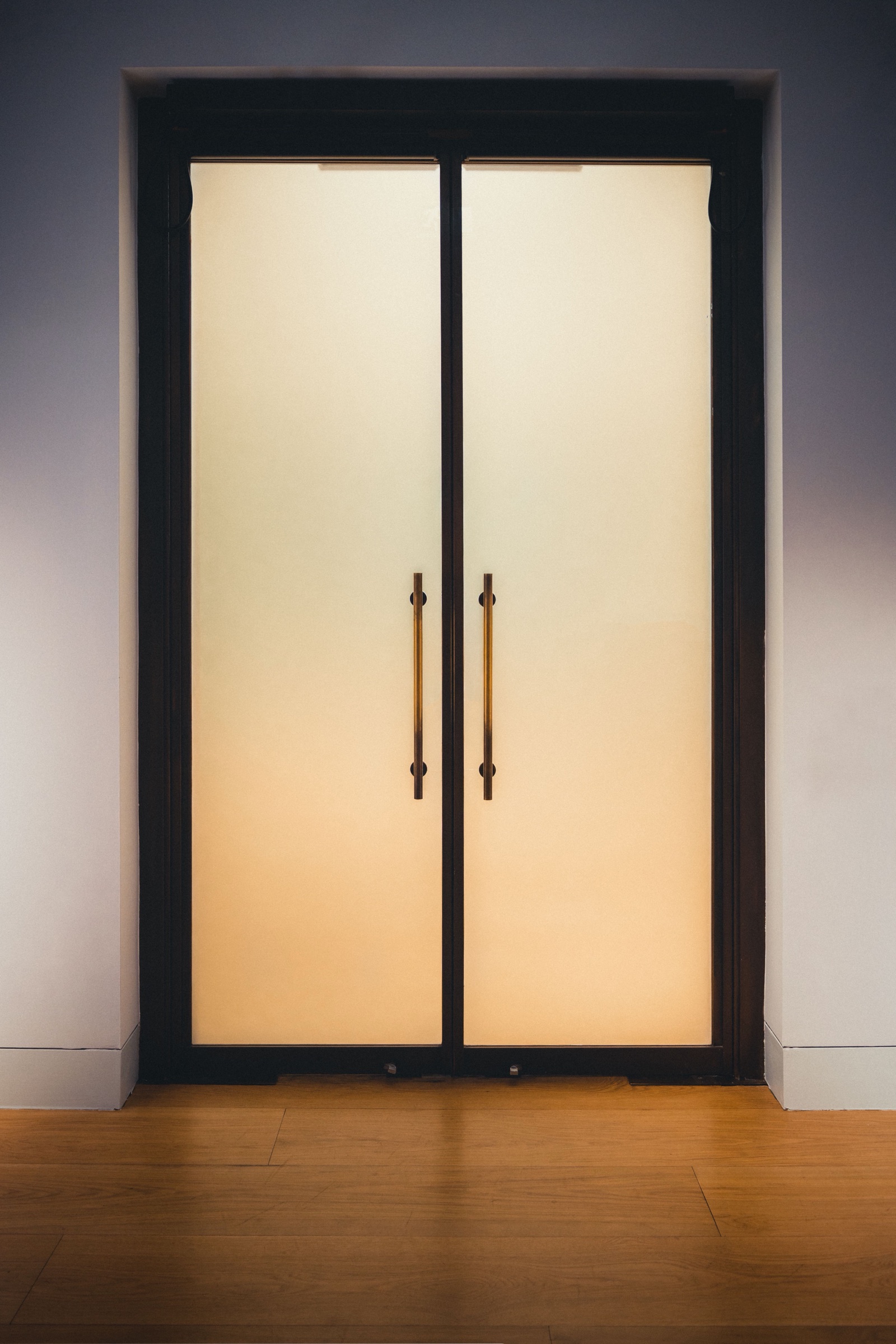A pair of full-height doors with frosted glass panels in slim dark frames and long brass bar handles, lit warmly from behind so the glass glows, set in a white wall above a wooden floor.