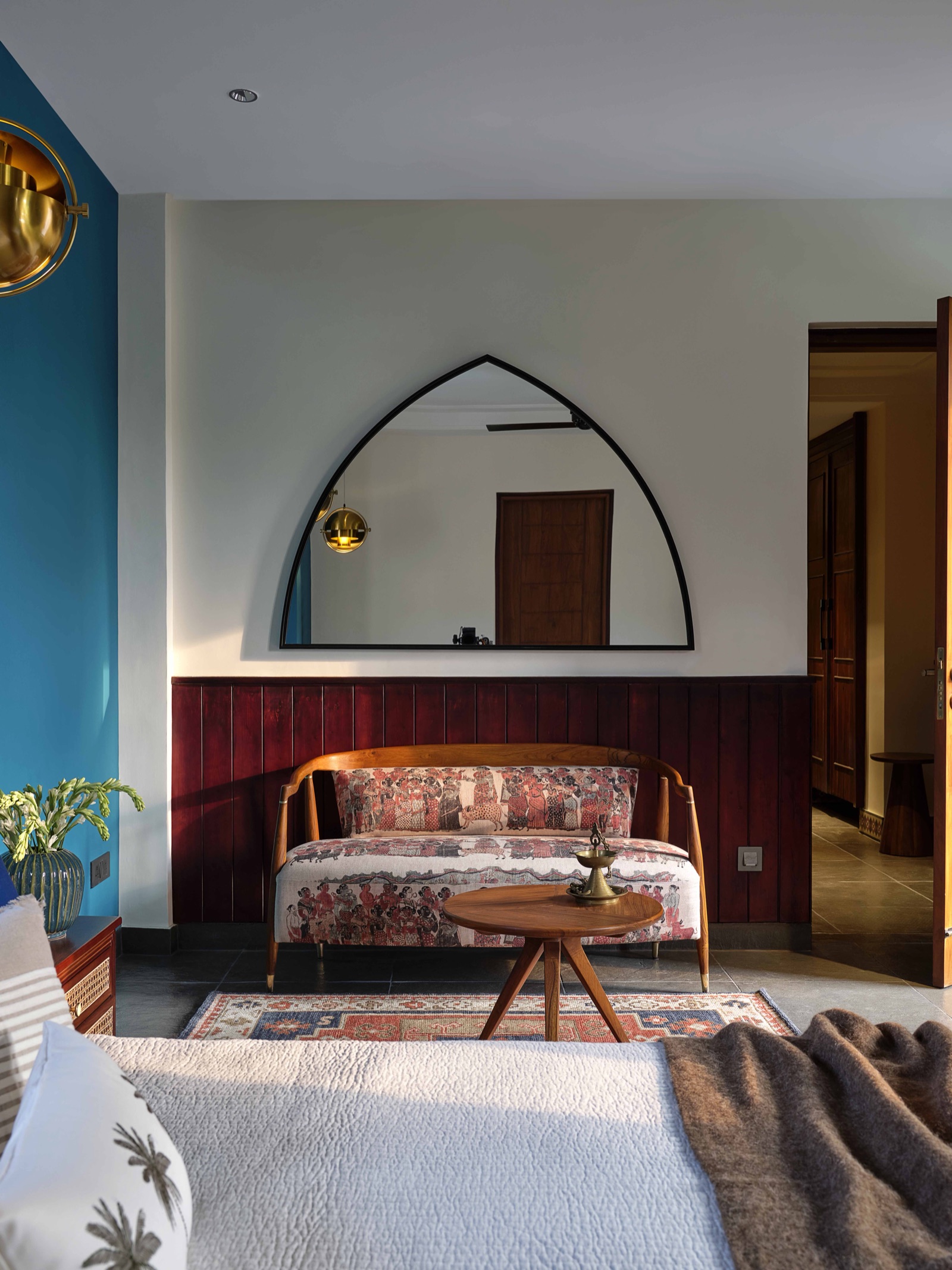 Bedroom corner with a teal blue wall on the left, an off-white wall with a large arched black-framed mirror, and deep maroon vertical timber panelling below it behind a printed wooden bench