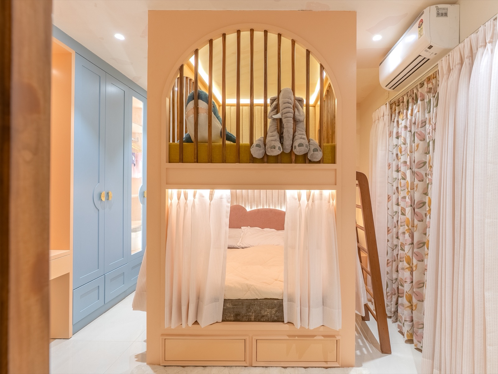 A child's bedroom with a powder-blue wardrobe on the left wall, its shaker-style shutters fitted with round gold handles and drawers below, beside a peach bunk bed with an arched, slatted upper level and curtains on the lower bed.