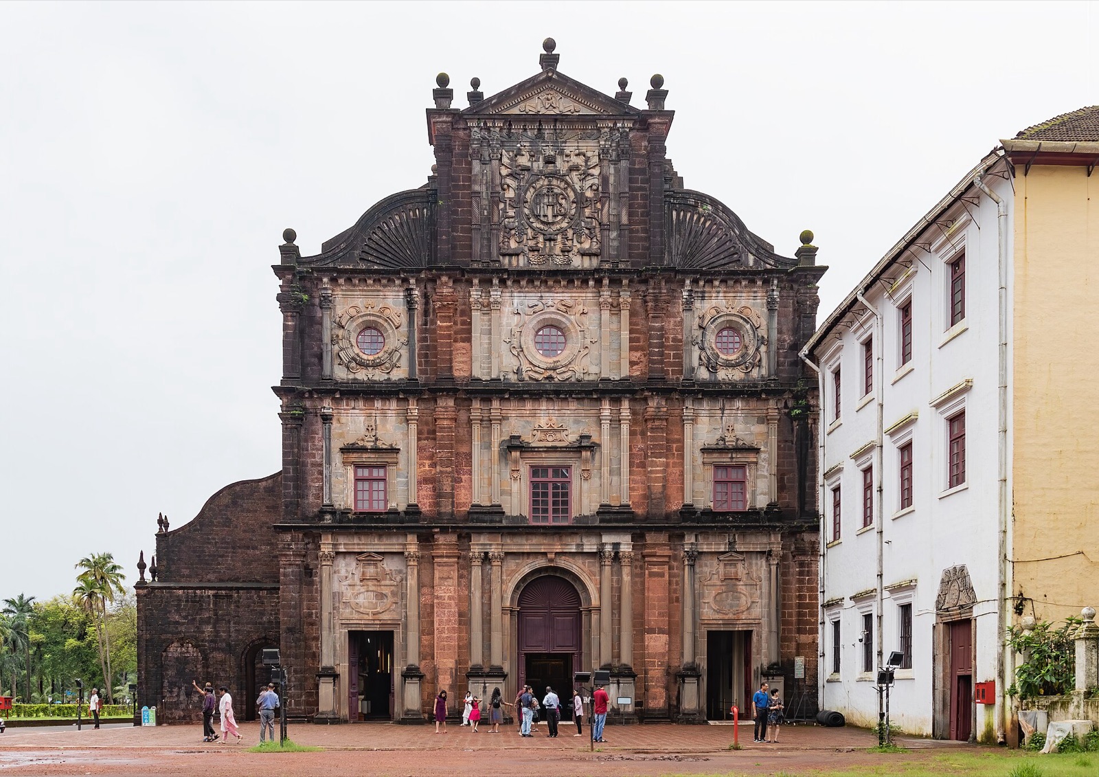 Front elevation of the Basilica of Bom Jesus in Old Goa, a three-storey Baroque facade of exposed red-brown laterite blocks framed by lighter grey carved stone pilasters, round windows and a tall central gable, photographed straight on under an overcast sky with a few visitors in the forecourt.