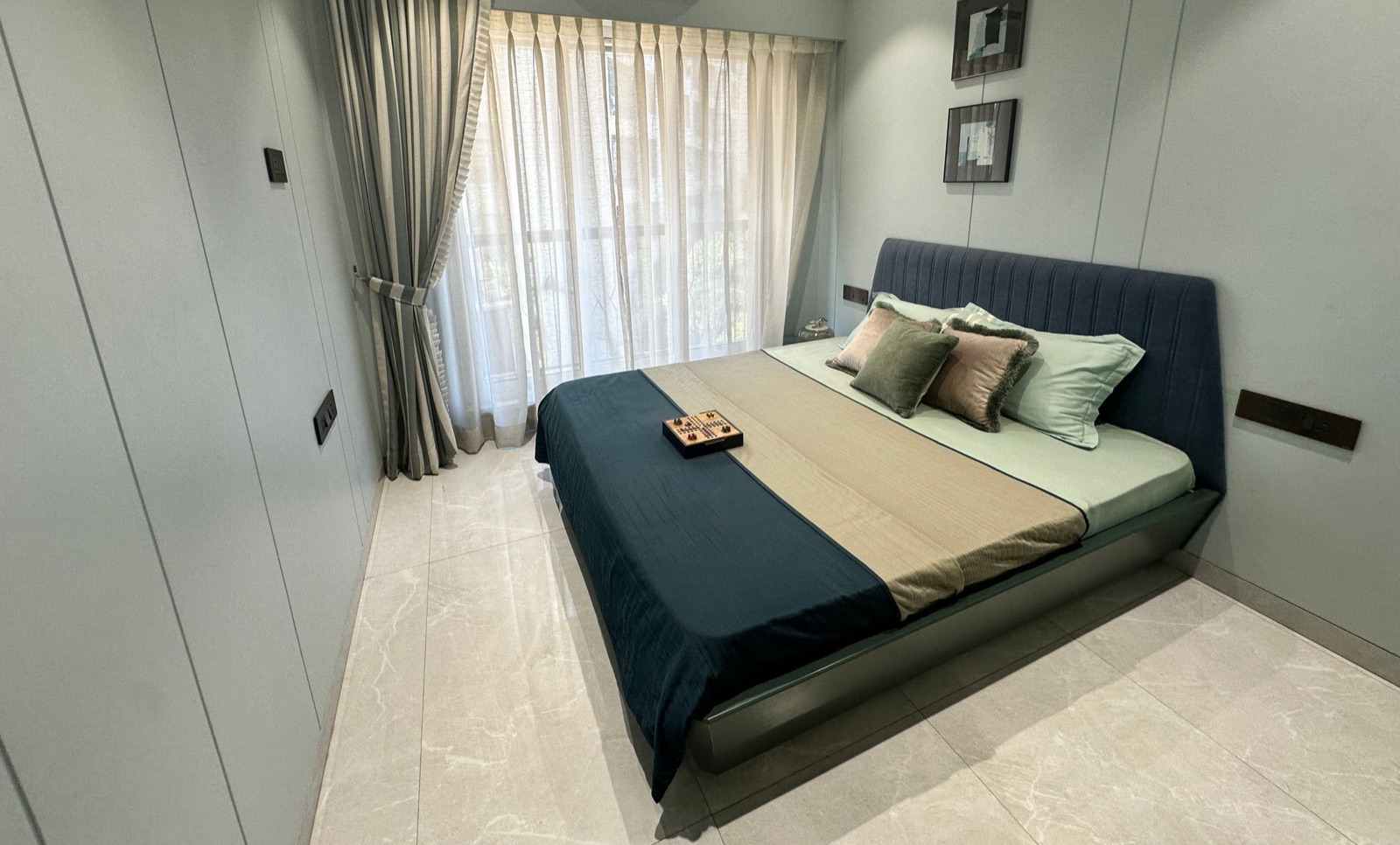 A compact Mumbai flat bedroom in pale grey-green: flush full-height wardrobe shutters painted the same colour as the walls on the left, a navy upholstered bed with no bedside tables, slim switch plates set into the wall beside the headboard, and sheer and striped curtains drawn back from a window