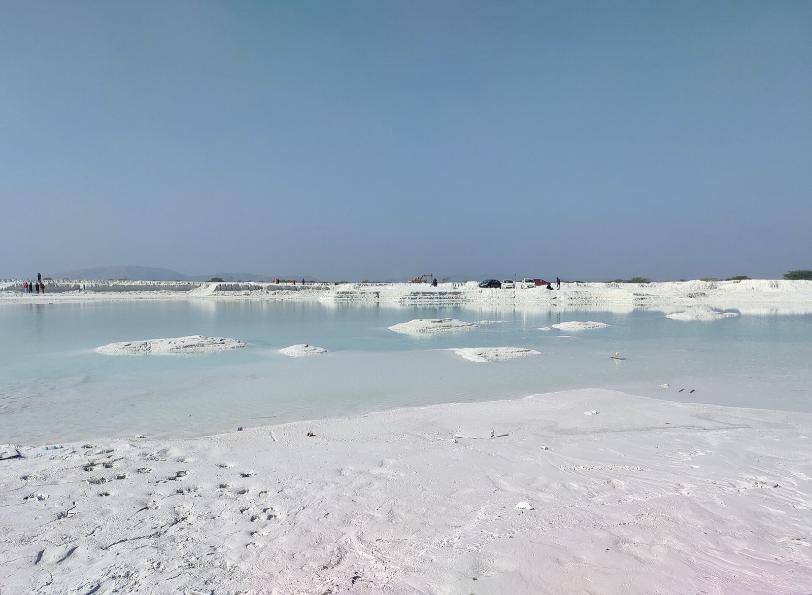 Flat white ground of dried marble slurry around a shallow pale blue pool, with low white mounds in the water, a long white embankment behind with a few small parked cars and visitors far away, and a hazy hill on the horizon