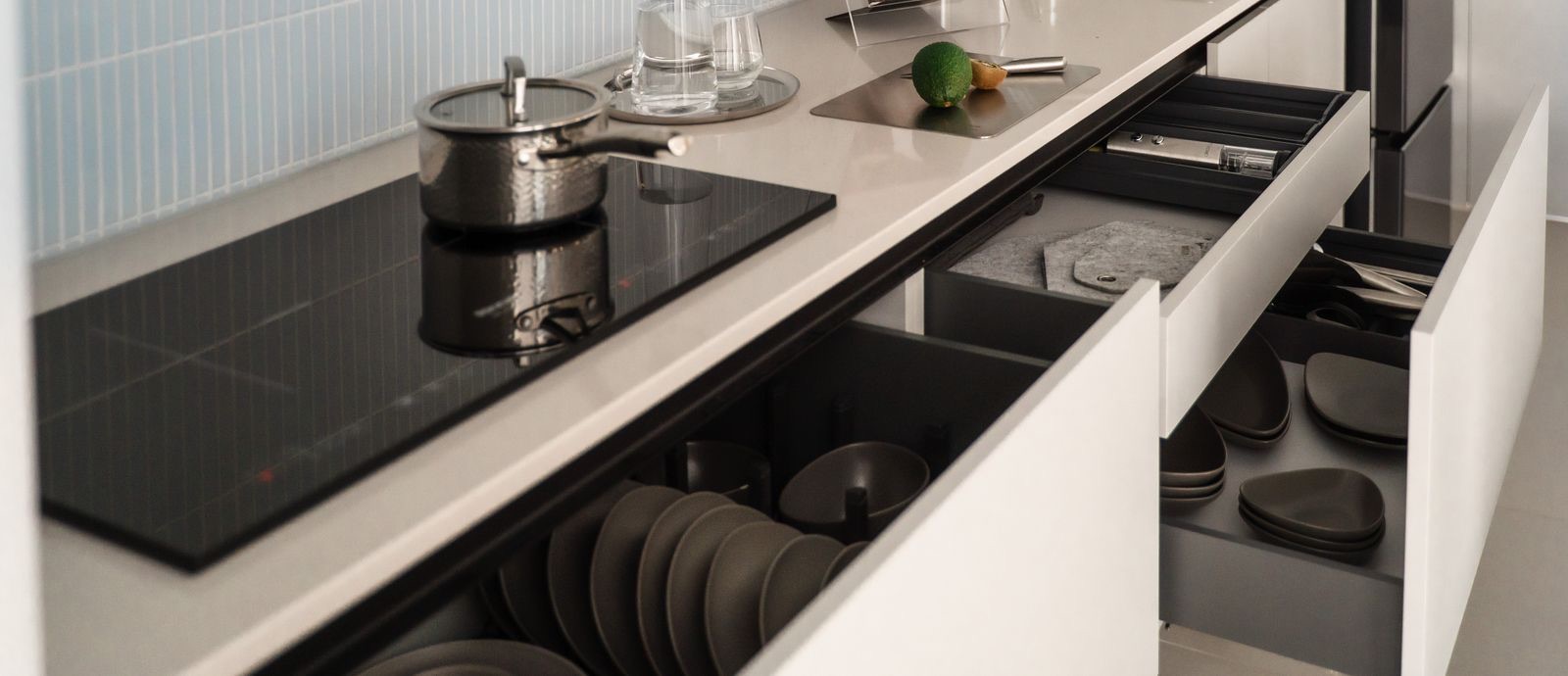 Base units under an induction hob with two wide drawers pulled out: the lower one holds plates upright between pegs, the upper one has a cutlery insert and stacked bowls
