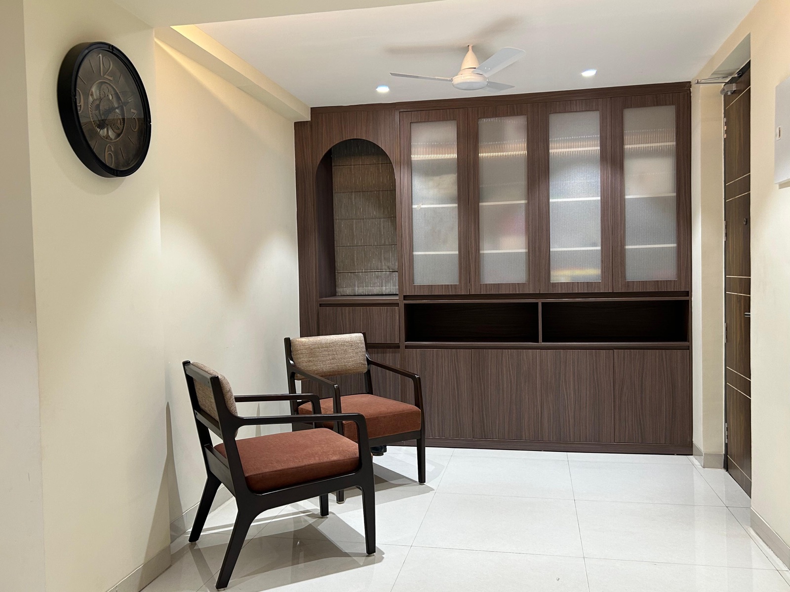 Entrance area of a Raipur 3BHK apartment with a full-height wood-finish storage wall: frosted glass shutters above, open shelves, an arched niche and closed cabinets below, with two upholstered armchairs on a white tiled floor