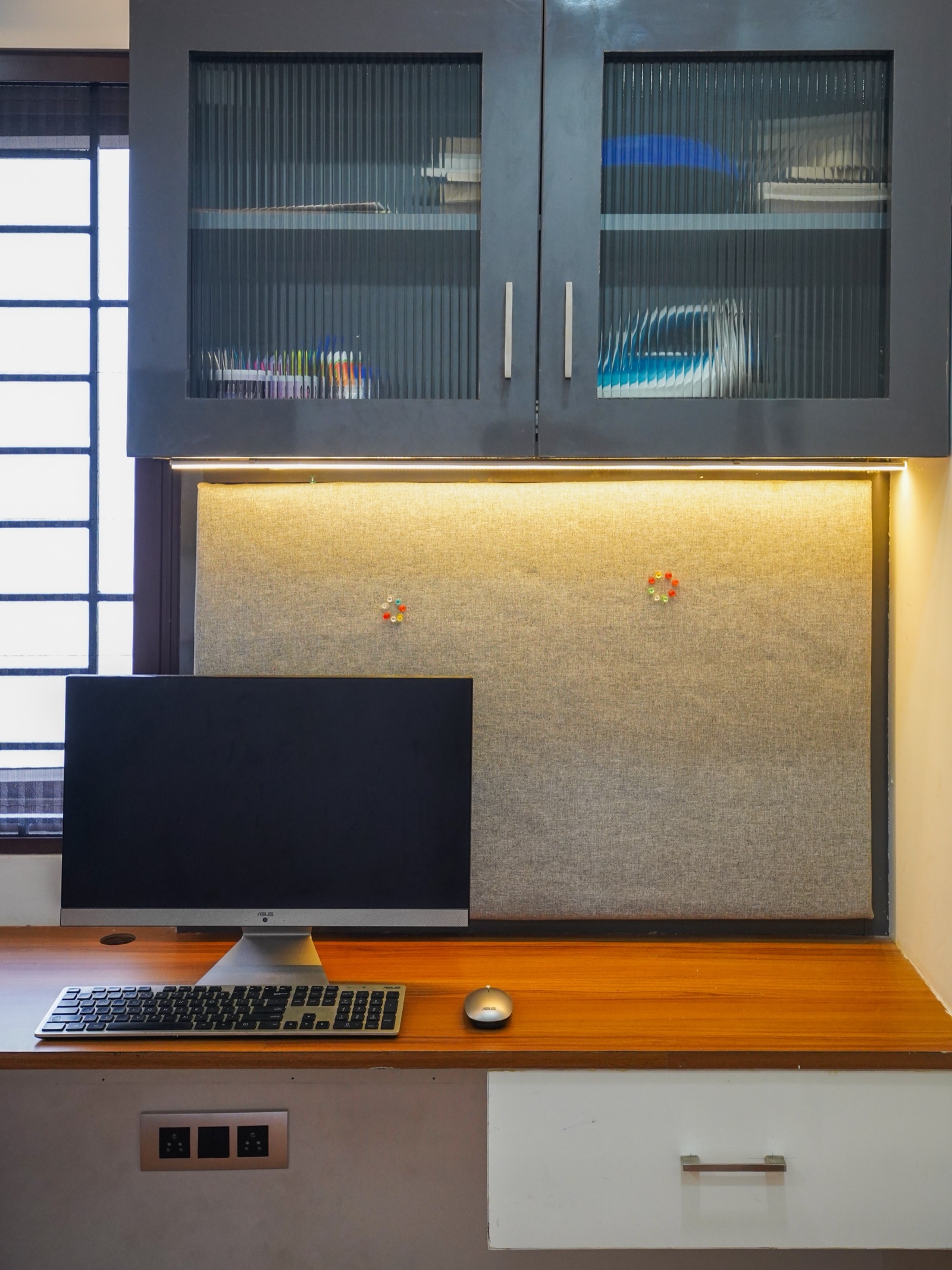 Built-in study desk in a Chennai home: a wood-finish worktop with a white drawer, a fabric pin-up board lit by a strip light, and grey overhead cabinets with fluted glass shutters beside a window