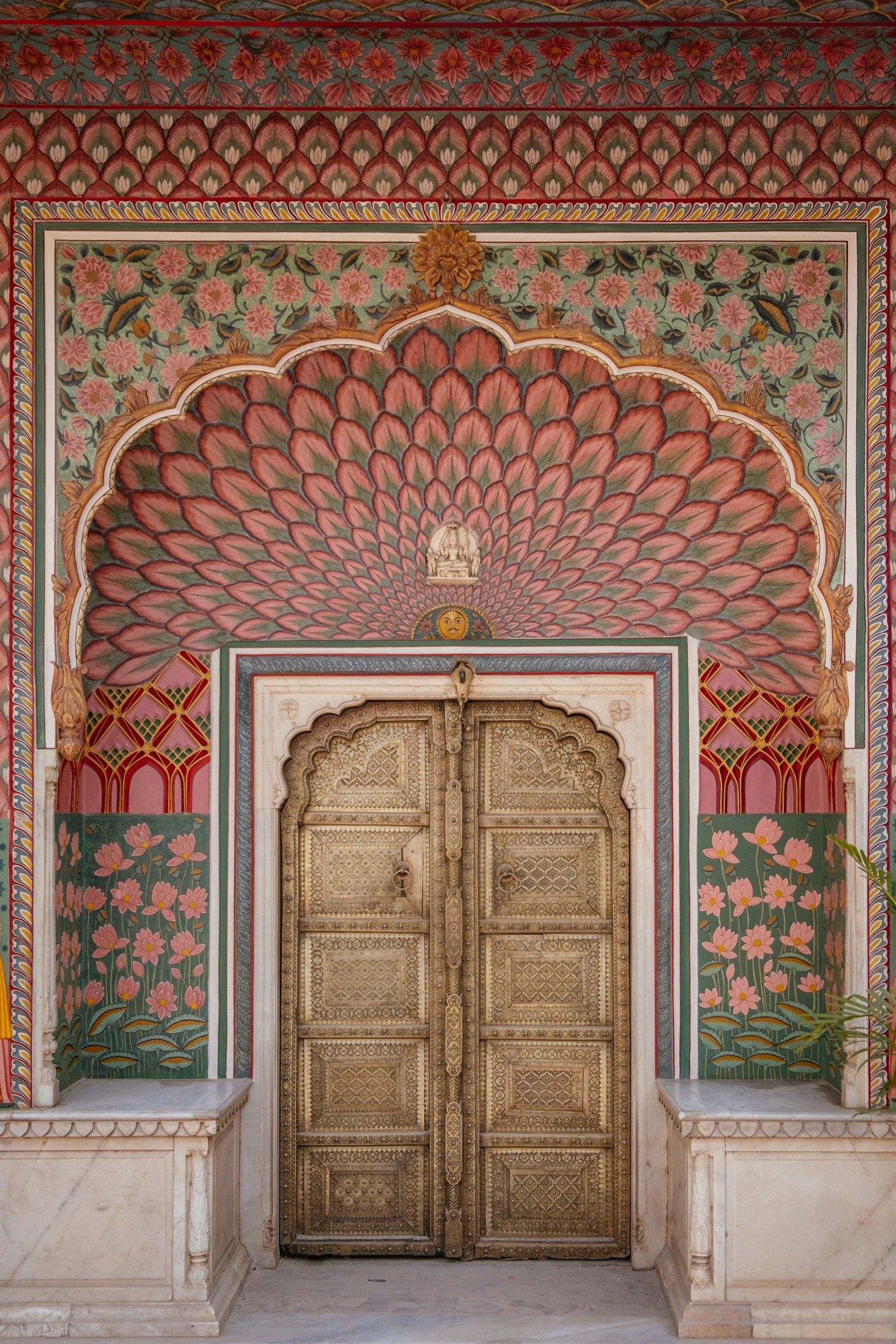 An ornate double door with raised metal panels set in a cusped arch, framed by painted pink and green lotus-petal and floral motifs and white marble plinths, at the City Palace in Jaipur's walled city