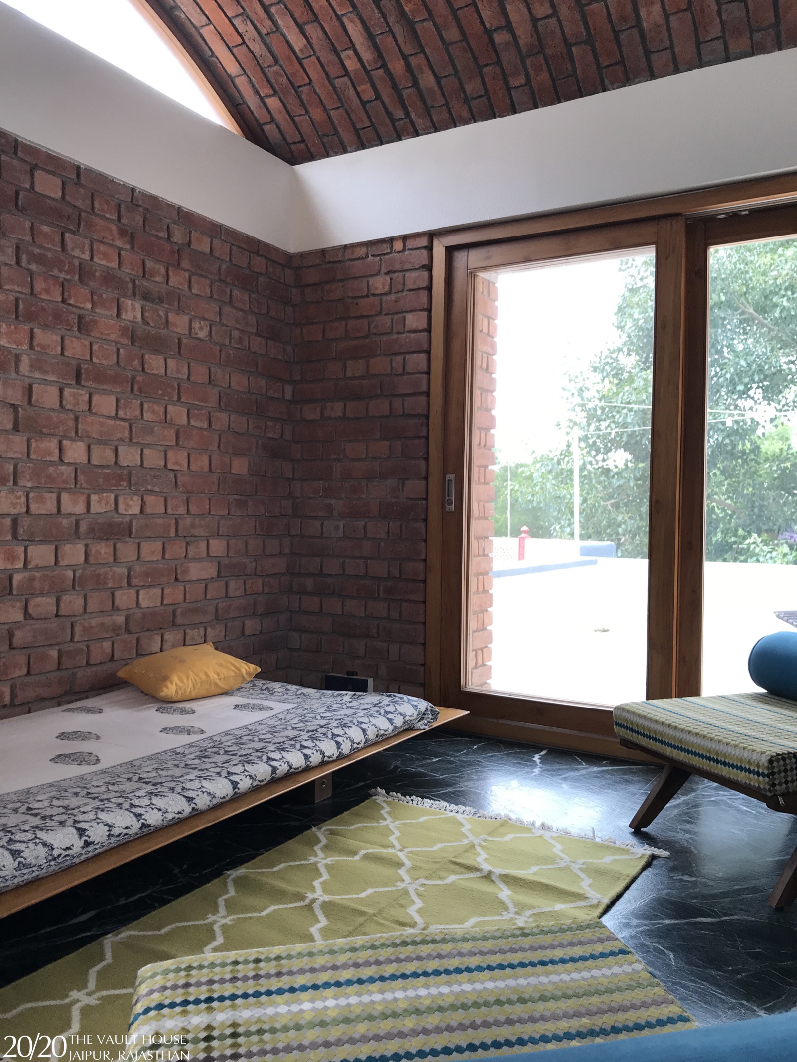 Corner of a Jaipur lounge with exposed brick walls under a brick vault, a timber-framed sliding glass door to a terrace, a low timber daybed with a printed cover and a green patterned rug on a dark marble floor