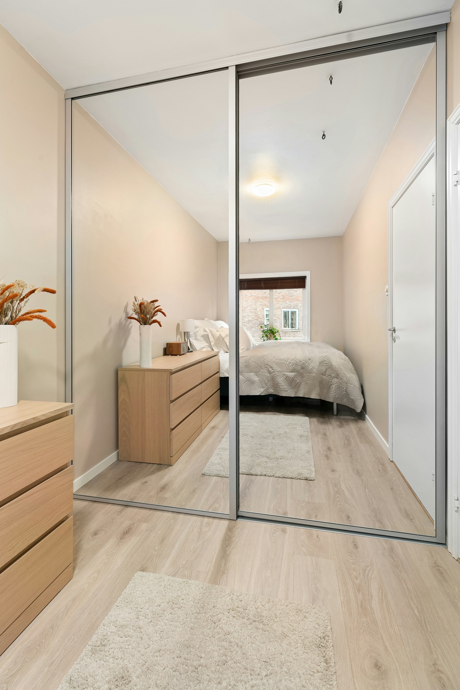 A two-panel sliding wardrobe with full-height mirror doors in thin aluminium frames, reflecting a narrow bedroom with a light oak chest of drawers, a dried-flower vase, a made bed and a window at the far end, on pale wood flooring.