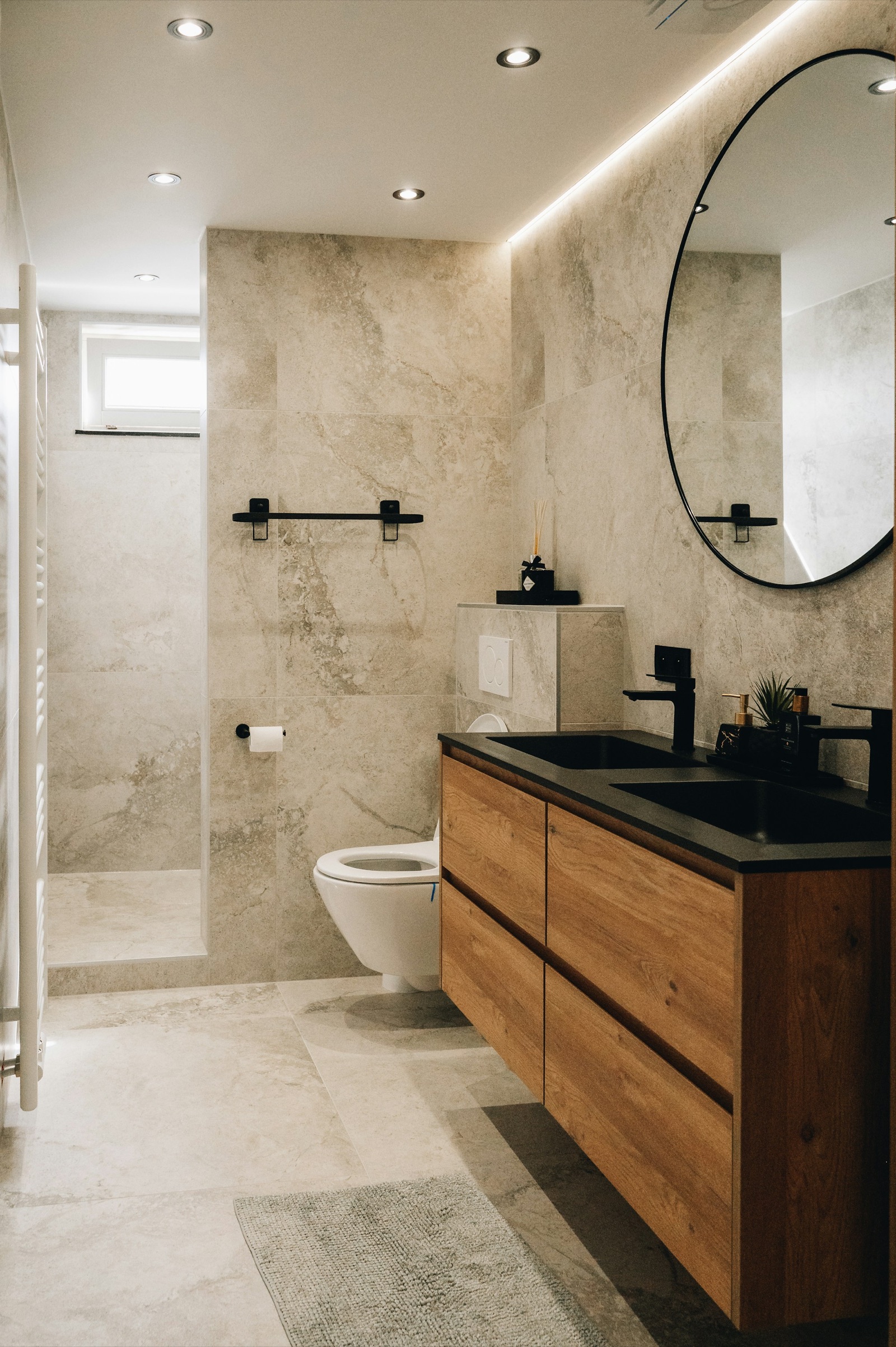 Bathroom finished in large stone-look porcelain tiles on walls and floor: a wall-hung oak double vanity with a black top and black mixers, a round black-framed mirror under a strip of cove light, a wall-hung WC with a flush plate on a tiled ledge, and a walk-in shower behind a tiled wall with a small high window