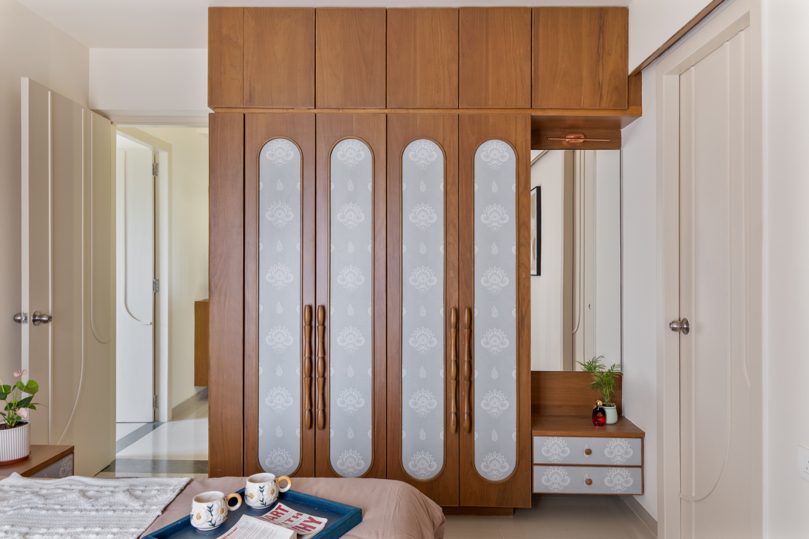 A full-height wooden wardrobe with four doors, each inset with a tall rounded panel of grey block-printed fabric and turned wooden handles, loft cabinets above, and a small dresser with a mirror and two drawers in a niche beside it.
