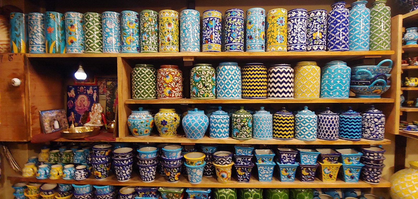Wooden shelves stacked with hand-painted Jaipur blue pottery: cylindrical jars, lidded pots, planters, mugs and bowls in cobalt blue, turquoise, yellow and green floral and chevron patterns