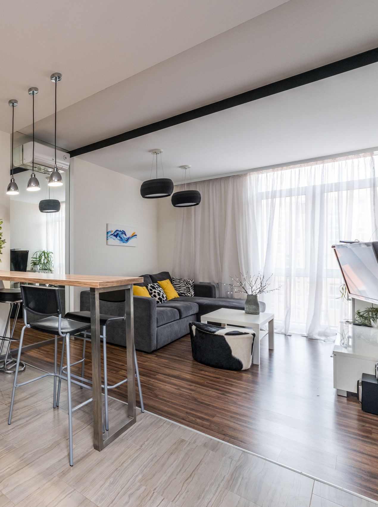 Open kitchen meeting a living room: a wood-topped bar counter on a steel frame with black stools stands on light stone-look floor tiles, which stop in a straight line where dark wood-look flooring starts under the grey sofa and coffee table.