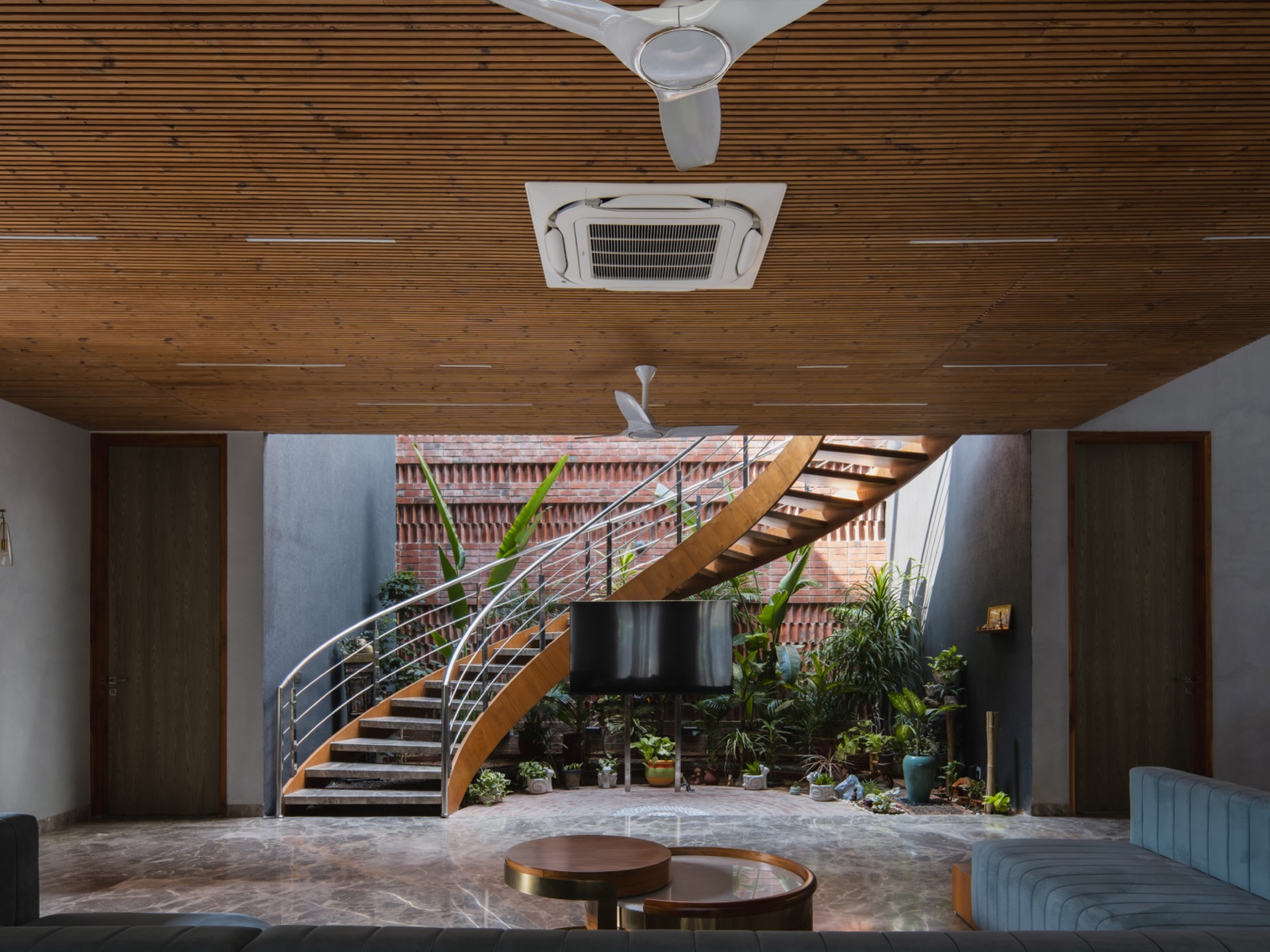 Living room with a flat ceiling of fine horizontal wood slats, slim linear light slots, a ceiling-mounted cassette air conditioner and two white ceiling fans. Beyond it, a curved timber staircase rises through an indoor courtyard of potted plants against a red brick jaali wall.