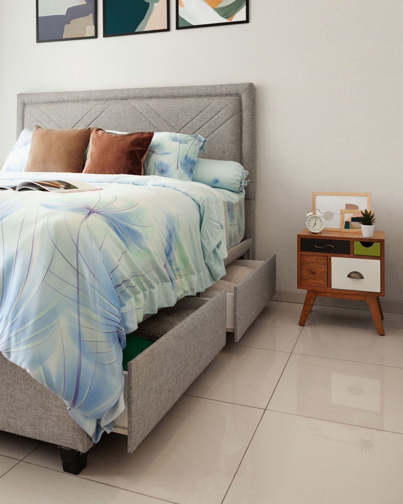 Grey upholstered bed with a stitched chevron headboard and two fabric-fronted drawers pulled open along its side, folded blankets inside, beside a small wooden bedside chest on a white tiled floor