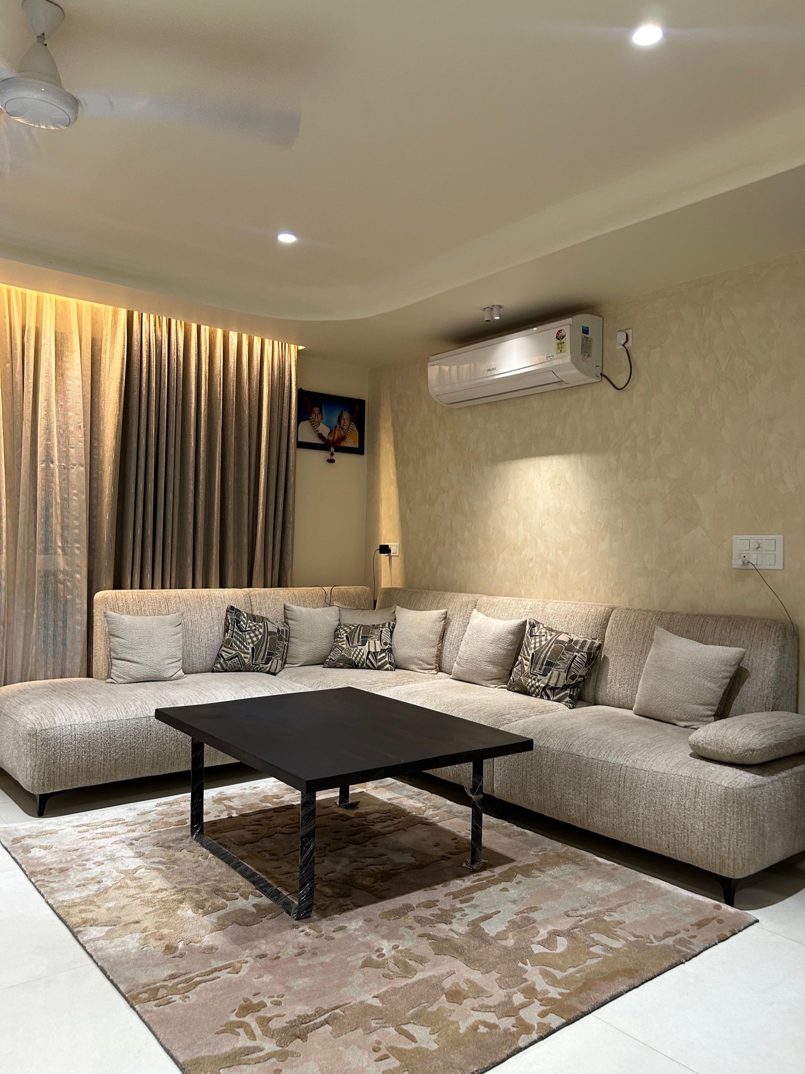 Living room in a Raipur apartment with a simple false ceiling: a shallow curved drop along the wall edge, two recessed downlights, a white ceiling fan in the plain centre, warm light washing the curtains, and a beige L-shaped sofa with a black coffee table on a patterned rug.