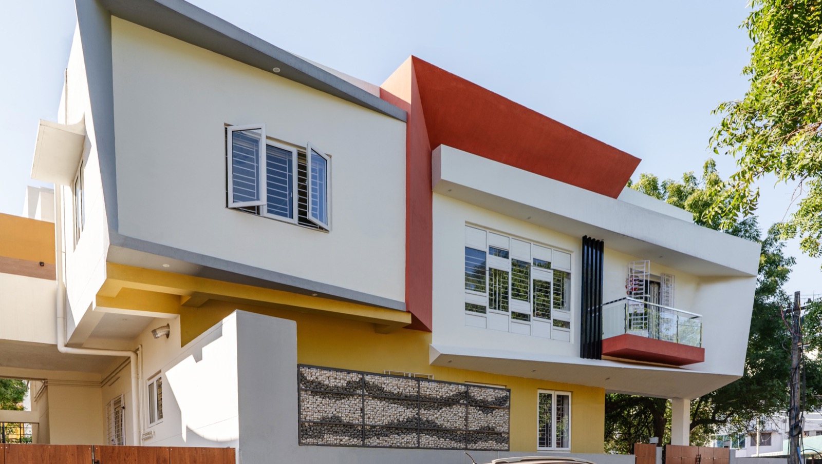 A two-storey white house with a red-brown angled fin, a yellow ground floor and deep projecting concrete frames over its windows, against a clear blue sky. A first-floor balcony has a glass railing on a red slab, and a gabion-style stone panel sits in the boundary wall beside wood-finish gates. A car is parked in front.